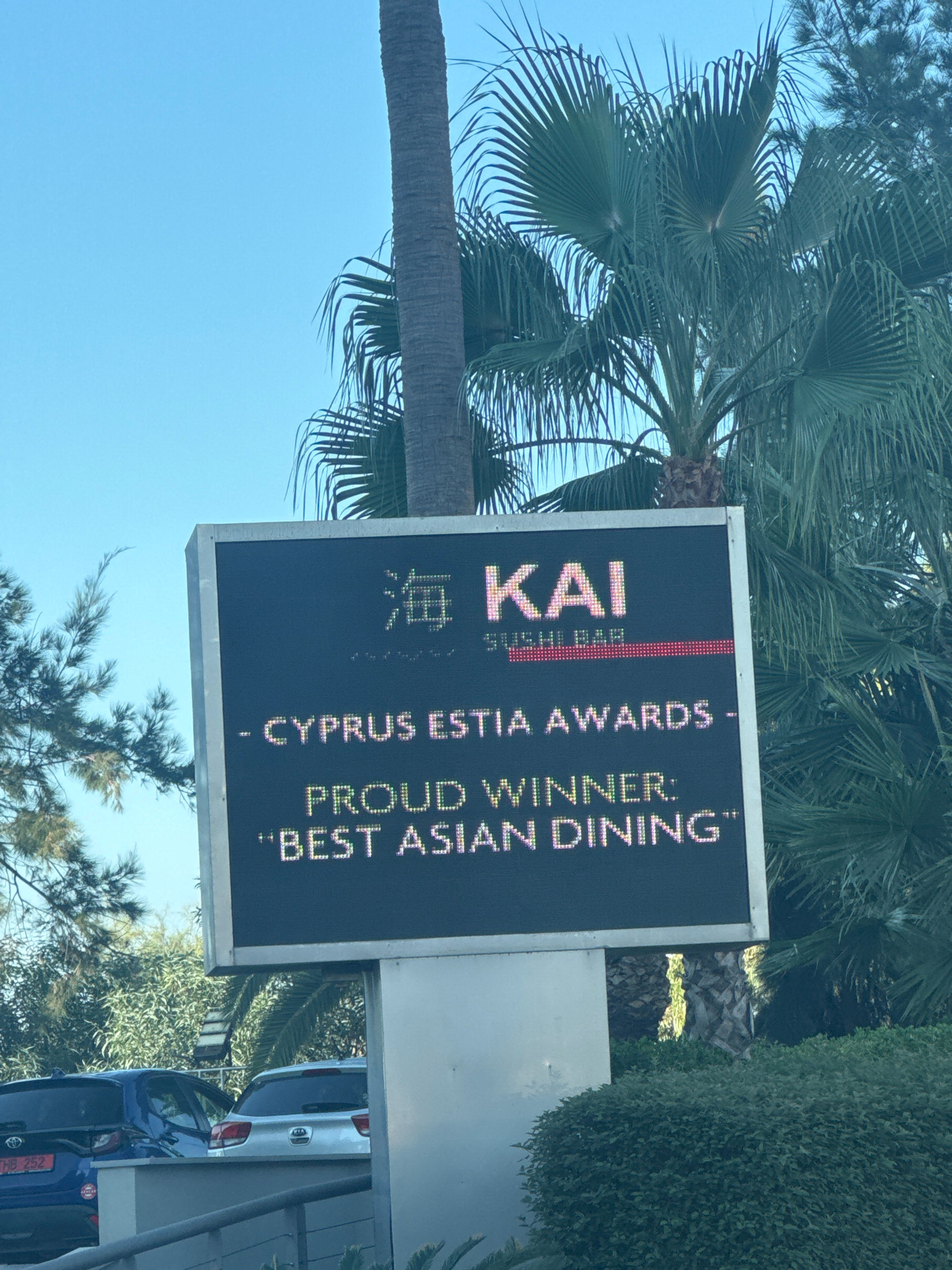 A digital sign displays "海 KAI SUSHI BAR" in large, illuminated text against a dark background. Below this, it reads "- CYPRUS ESTIA AWARDS - PROUD WINNER: "BEST ASIAN DINING"". The sign is mounted on a white pole and surrounded by palm trees and lush green foliage. In the foreground, the tops of two parked cars are visible. The sky is clear blue. The scene is outdoors during daylight. Based on the text on the sign, the location is Limassol, Cyprus, specifically at Kai Sushi Bar.