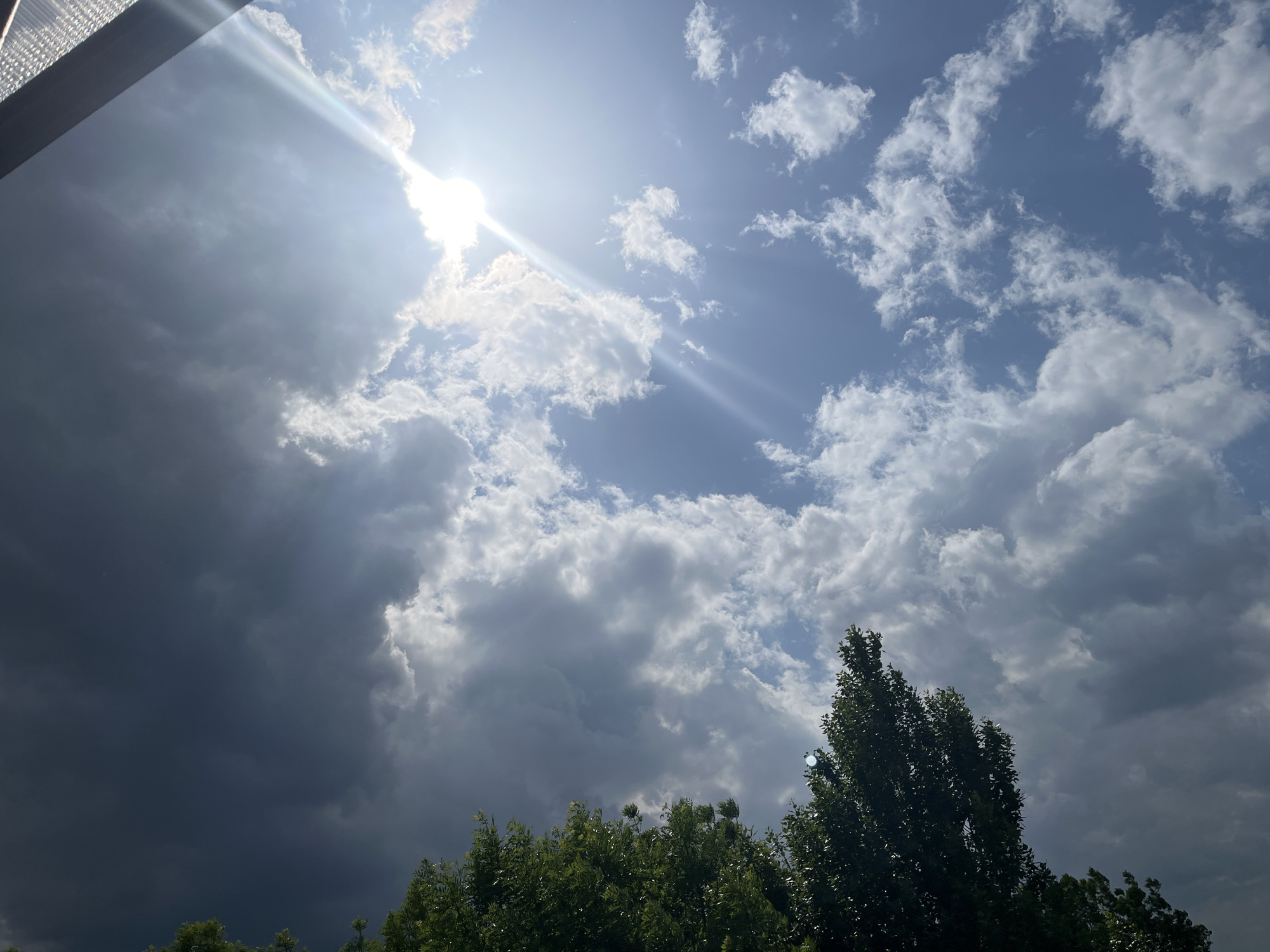 The image captures a sky with a mix of dark storm clouds and bright, sunlit clouds. The sun is visible, casting rays through a break in the clouds. The tops of green trees are visible in the lower portion of the image, partially obscured by the clouds. In the upper left corner, a portion of a building or structure is visible. The location is Unknown, Unknown.