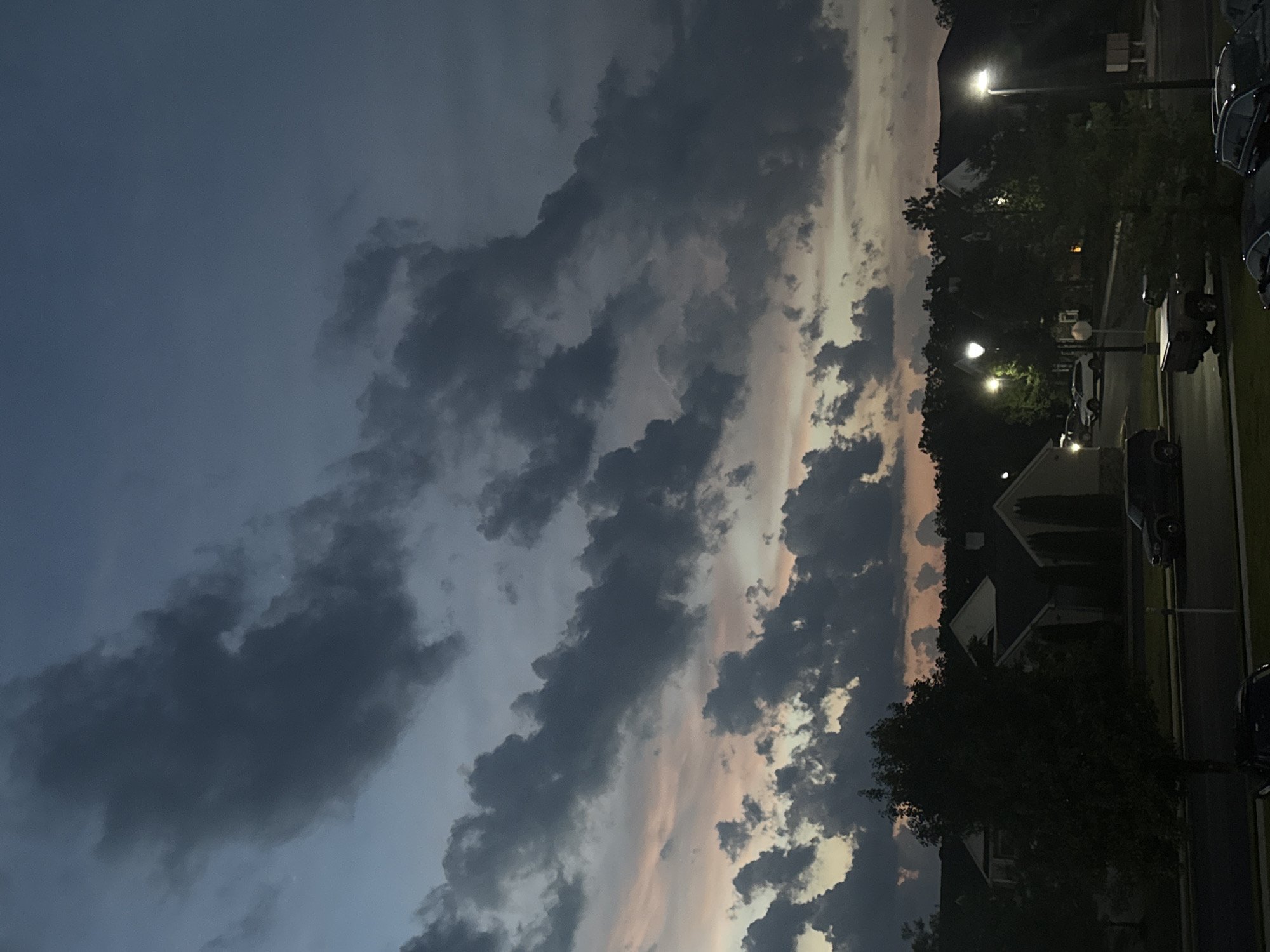 This image depicts a twilight scene over a residential area in Allen Township, United States of America. The sky is dominated by dramatic, dark clouds with hints of pink and orange from the setting sun peeking through. Below the sky, a line of trees partially obscures a row of houses with peaked roofs and some with chimneys. Several streetlights are illuminated, casting pools of light onto the street and sidewalks. Various vehicles are parked on the street and in what appear to be driveways, including a dark-colored SUV, a pickup truck, and several other cars. The overall atmosphere is calm and still, characteristic of the early evening.