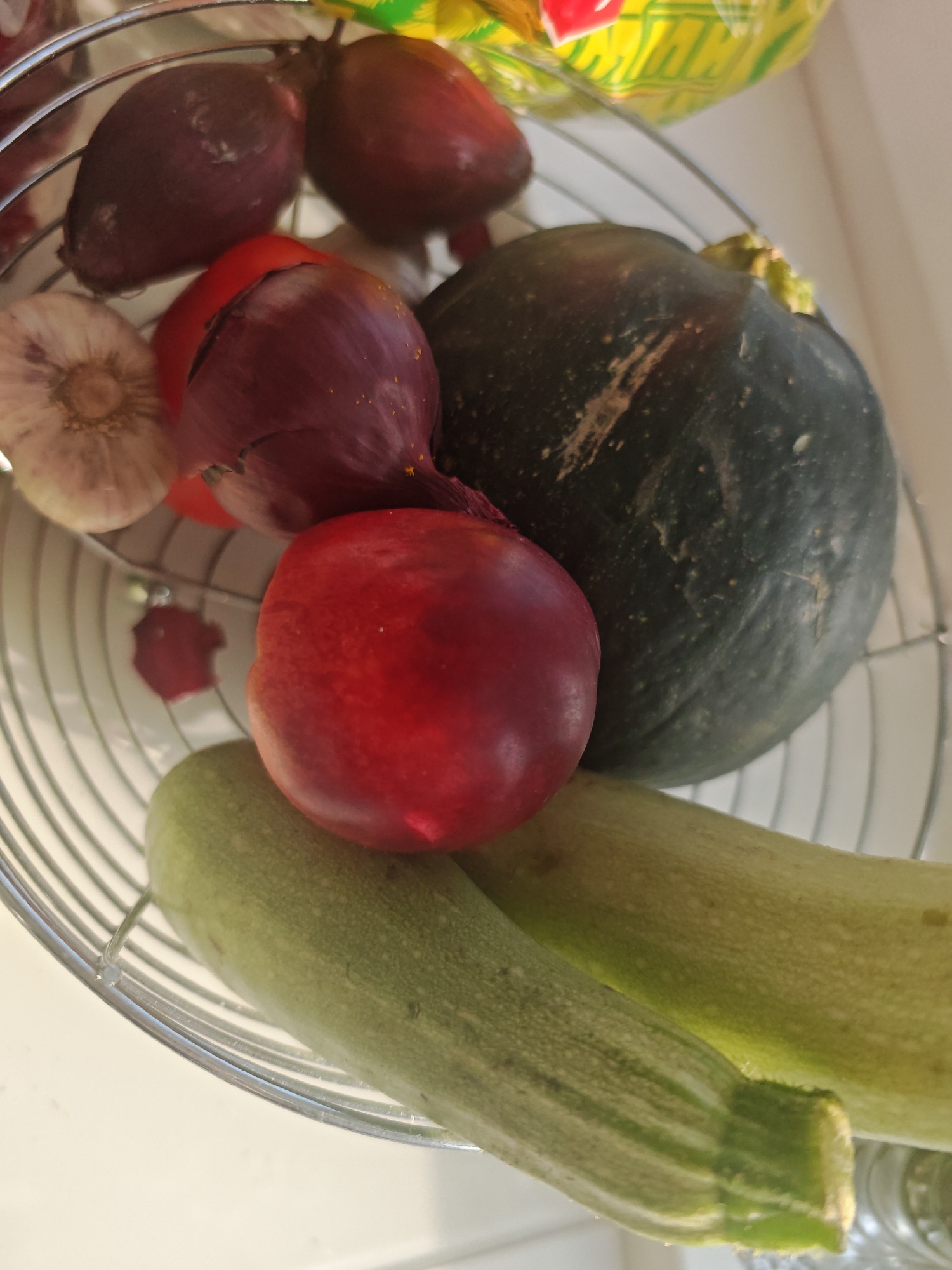 The image shows a close-up, overhead view of a metal basket filled with various produce. The basket contains two light green zucchini, a dark green round squash, a red nectarine, a head of garlic, and two red onions, one of which has a tomato partially visible behind it. The basket is set against a plain white surface. The lighting is bright, suggesting an indoor environment. There are no people or animals visible in the image. The scene appears to be a still life arrangement of fresh vegetables and fruit. The specific location of Riga, Latvia, cannot be determined from the visual information provided in the image.