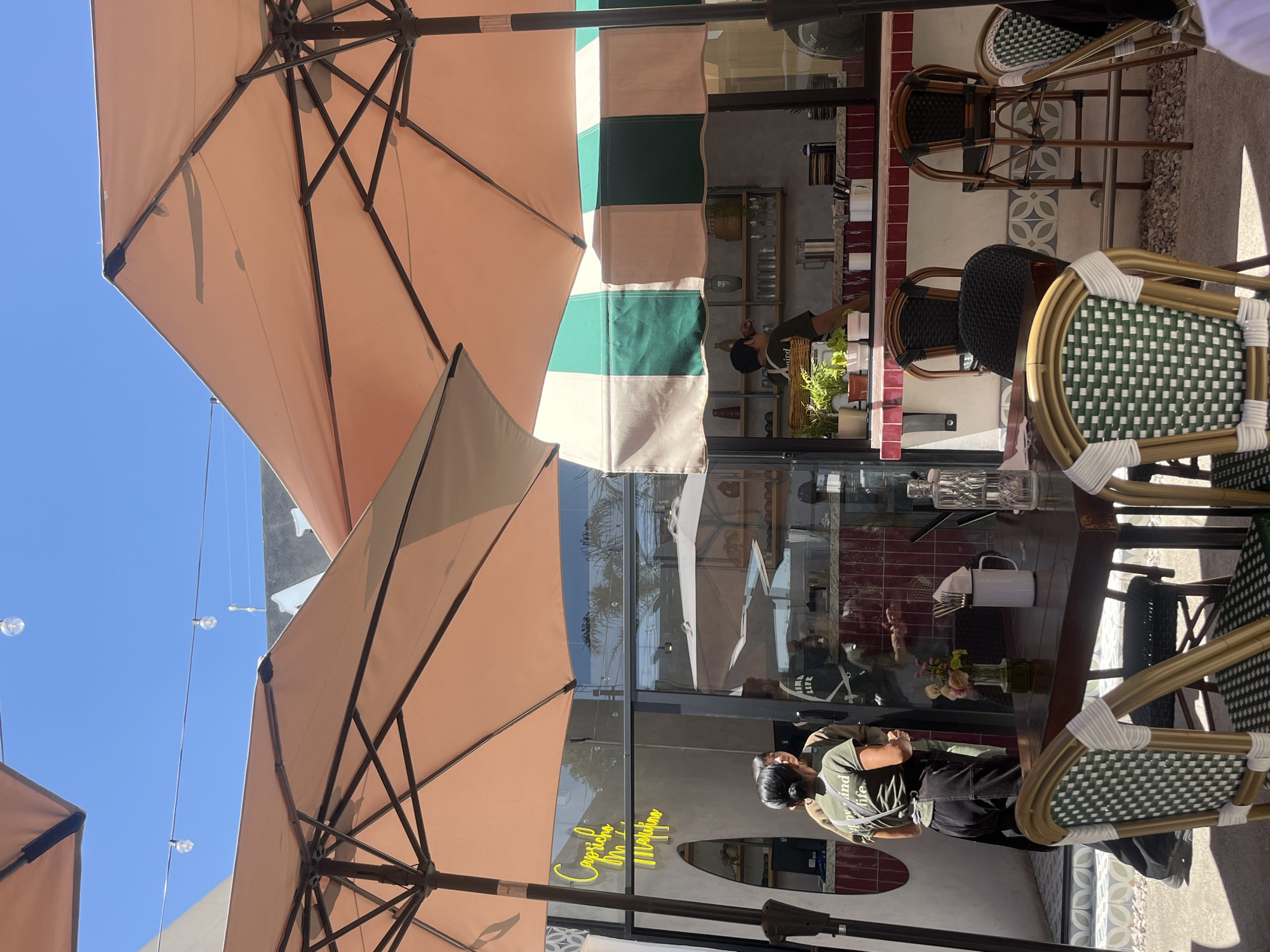The image shows an outdoor seating area of a cafe or restaurant in San Bernardino Tlaxcalancingo, Mexico. The scene is taken during the daytime with bright sunlight. Two large, open peach-colored umbrellas dominate the upper part of the frame, providing shade to the tables.
In the background, a storefront with glass windows reflects the umbrellas and the clear blue sky above. A sign with neon yellow lettering, visible through the glass, spells out "caprichos On & Off", presumably the name of the establishment.
Several chairs with green and white woven seats are arranged around a dark-stained wooden table. A person wearing a dark apron and light green shirt with printed text is visible near the table, appearing to be cleaning or setting it. Cutlery, a white mug, and a small vase with flowers are placed on the table. Other chairs and stools are seen in the background, close to the storefront. The flooring is a mix of tiling and gravel. A fabric awning with green and cream-colored blocks is partially visible. Overhead, strings of decorative lights hang above the seating area.