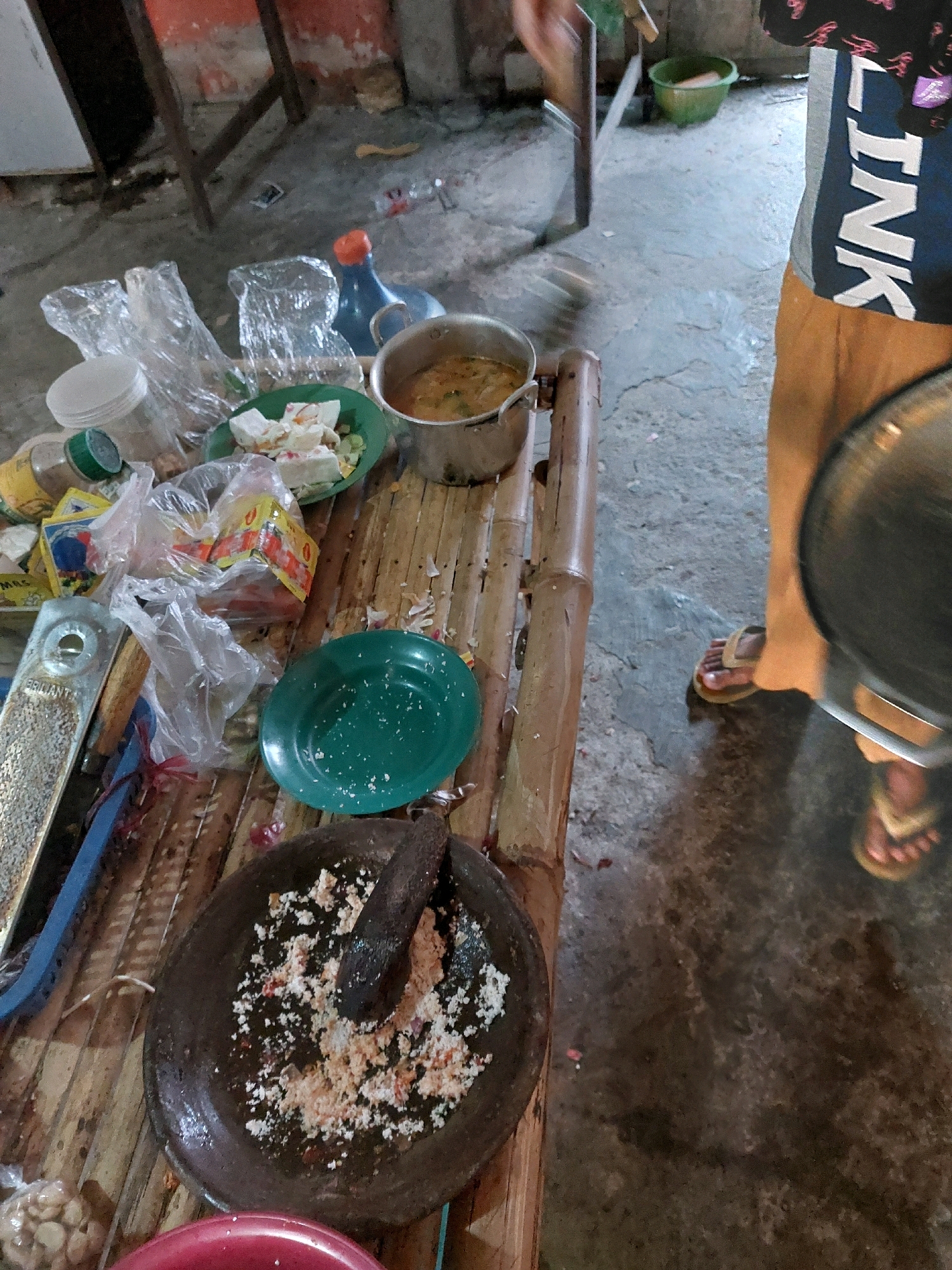 The image depicts an outdoor kitchen scene in Sarangan, Indonesia. A person, wearing orange pants and a black and white shirt with the word "LINK" prominently displayed, stands in the lower right of the frame, holding a large metal pot or pan. Scattered across a bamboo table are various kitchen items. These include plastic bags filled with what appear to be food items, small containers of spices or seasonings, a green plate with some food debris, a mortar and pestle with ground spices, and a metal grater. In the background, a blue plastic bottle with an orange cap is visible, along with a green bucket. The ground is made of concrete and appears wet in some areas. The overall impression is one of a functional, albeit somewhat cluttered, outdoor cooking area.