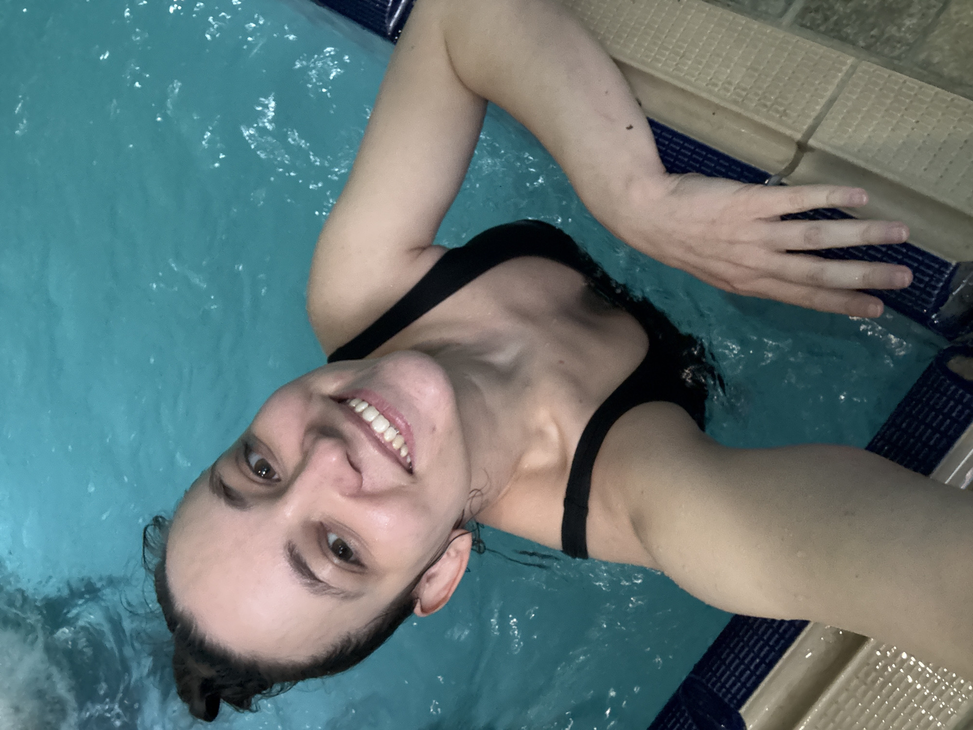 A woman is in a pool, holding onto the side. She is smiling and appears to be taking a selfie. She is wearing a black swimsuit. The pool is tiled with blue and white tiles. The location is most likely a swimming pool, possibly in the city of Chaikovsky, Russia.