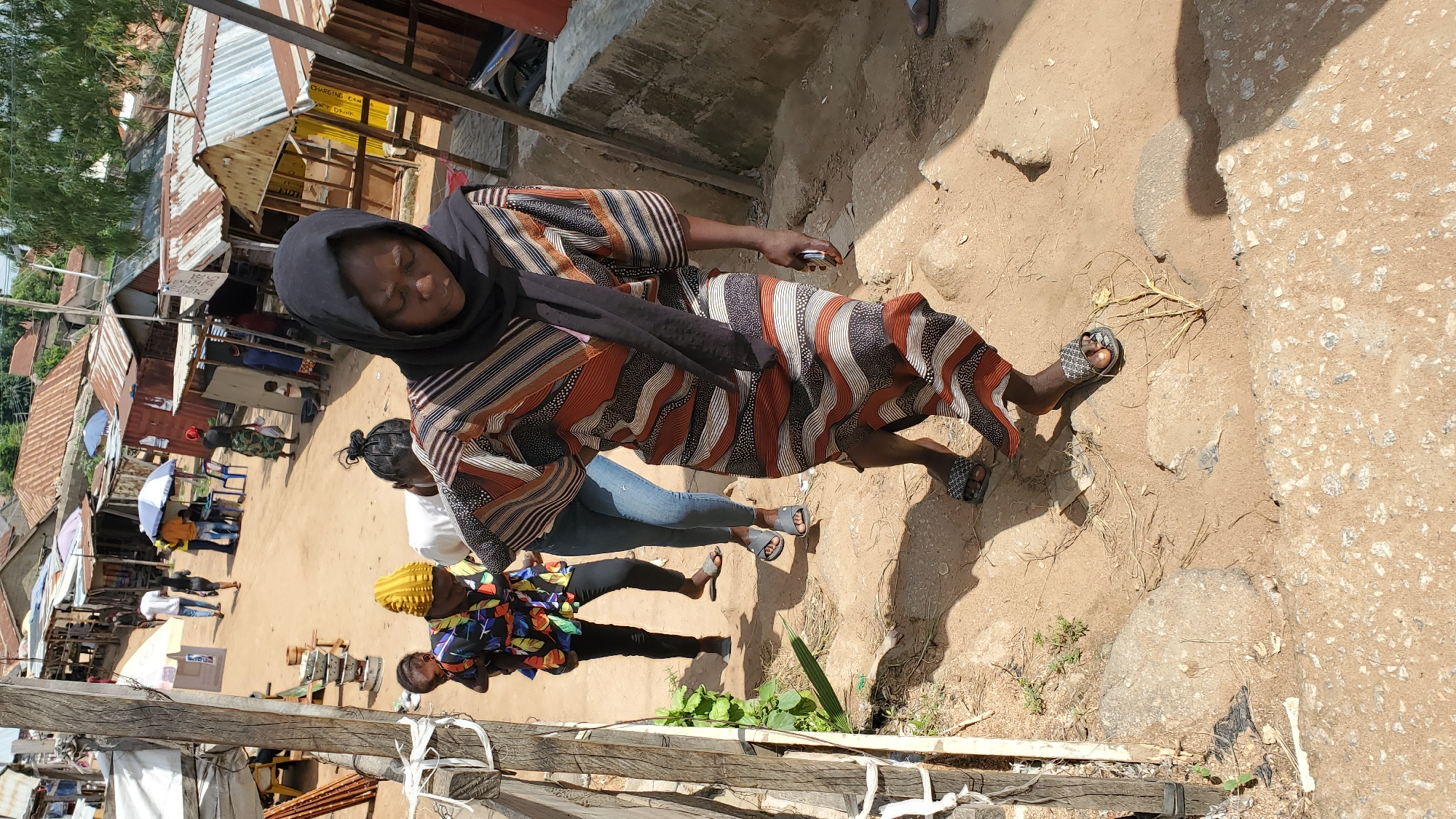In Jalingo, Nigeria, a group of individuals navigates a dusty, uneven pathway lined with makeshift structures. A woman in a patterned dress and black hijab leads the way, descending a dirt incline. Behind her, a woman in a colorful, multi-patterned top carries a baby, followed by another woman in blue jeans. The background reveals a bustling marketplace with stalls covered by tarps and corrugated metal roofs. The ground is a mixture of sand, dirt, and scattered debris, suggesting an unpaved urban environment. Sunlight casts strong shadows, indicating a bright day.