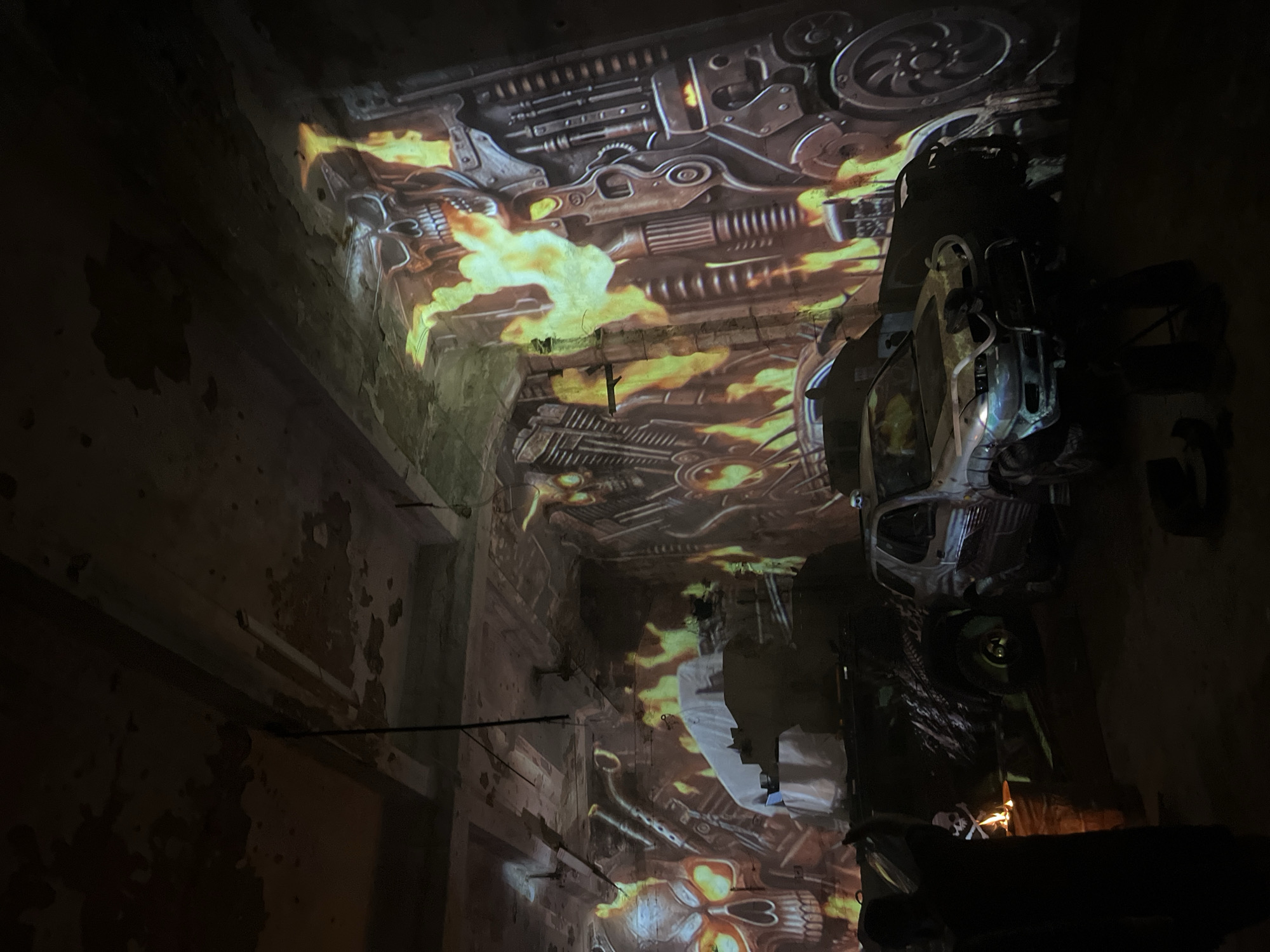 The image depicts a dimly lit, industrial-style interior space, possibly an abandoned building or a themed exhibit. The walls and ceiling are adorned with projected imagery featuring flaming skulls, mechanical components, and what appear to be stylized engine parts, creating a post-apocalyptic or heavy metal aesthetic. In the foreground, several vehicles are visible, including a white SUV that has been modified with a bull bar and other rugged accessories. The SUV is parked next to a darker, larger vehicle, possibly a truck or another SUV, also with a rough appearance. To the left, a person is silhouetted against a barrel with flames erupting from it, resembling a makeshift fire. The overall atmosphere is dark and dramatic, illuminated by the projected visuals and the fire. The location is stated to be in the city of Қарғалы, Kazakhstan.