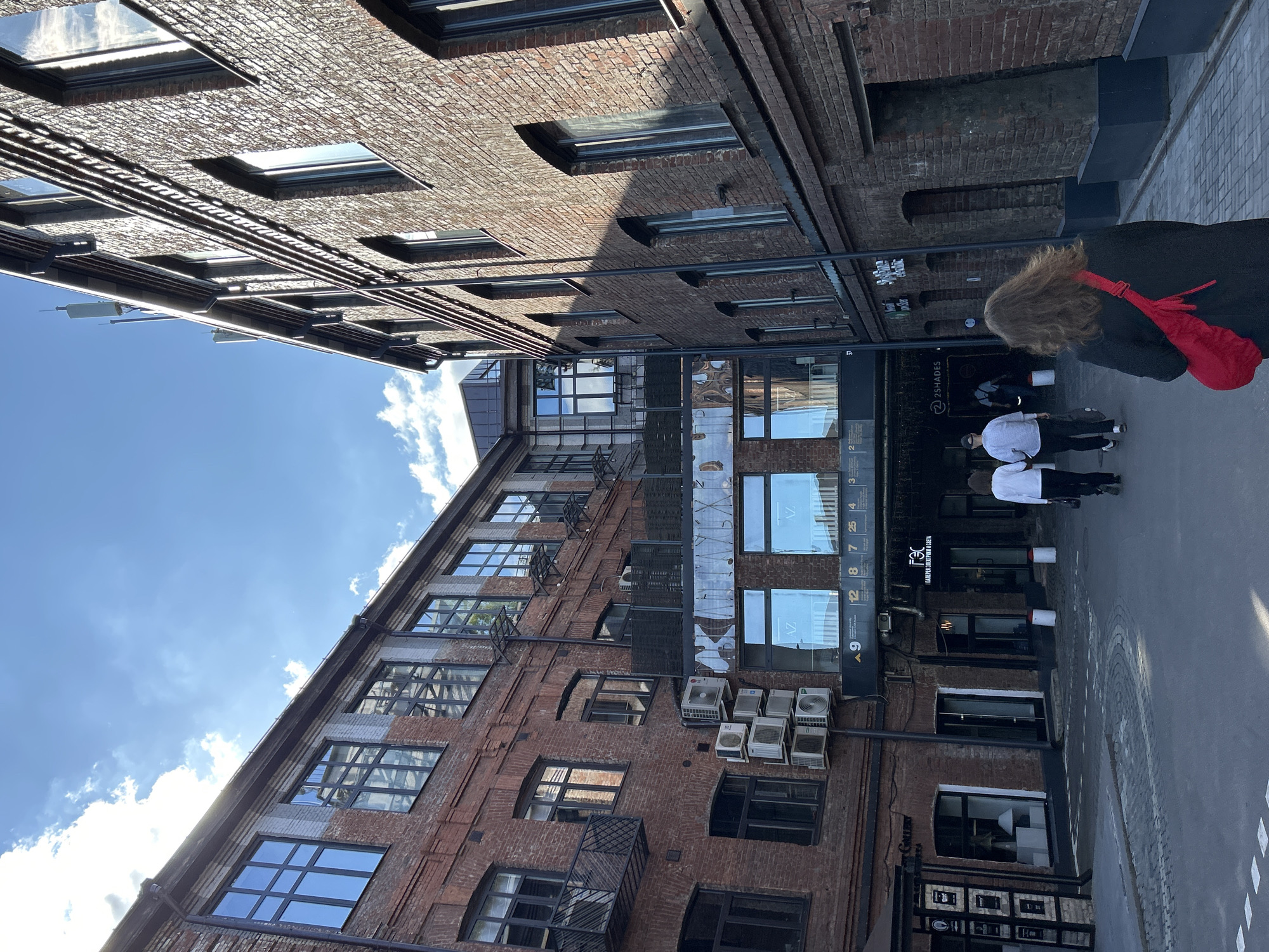 The image depicts an outdoor courtyard between two red brick buildings, under a bright blue sky with scattered clouds. The buildings have numerous large windows and some visible air conditioning units. In the foreground, a person with wavy brown hair wearing a black jacket and a red crossbody bag walks away from the camera. Two other individuals, one wearing a white long-sleeved shirt and black pants, and another in a light-colored long-sleeved shirt and black pants, are walking ahead in the same direction. Signage is visible on the buildings, including one with Russian text that appears to be a business name. The ground is paved with cobblestones. The overall scene suggests an urban environment with architectural interest.