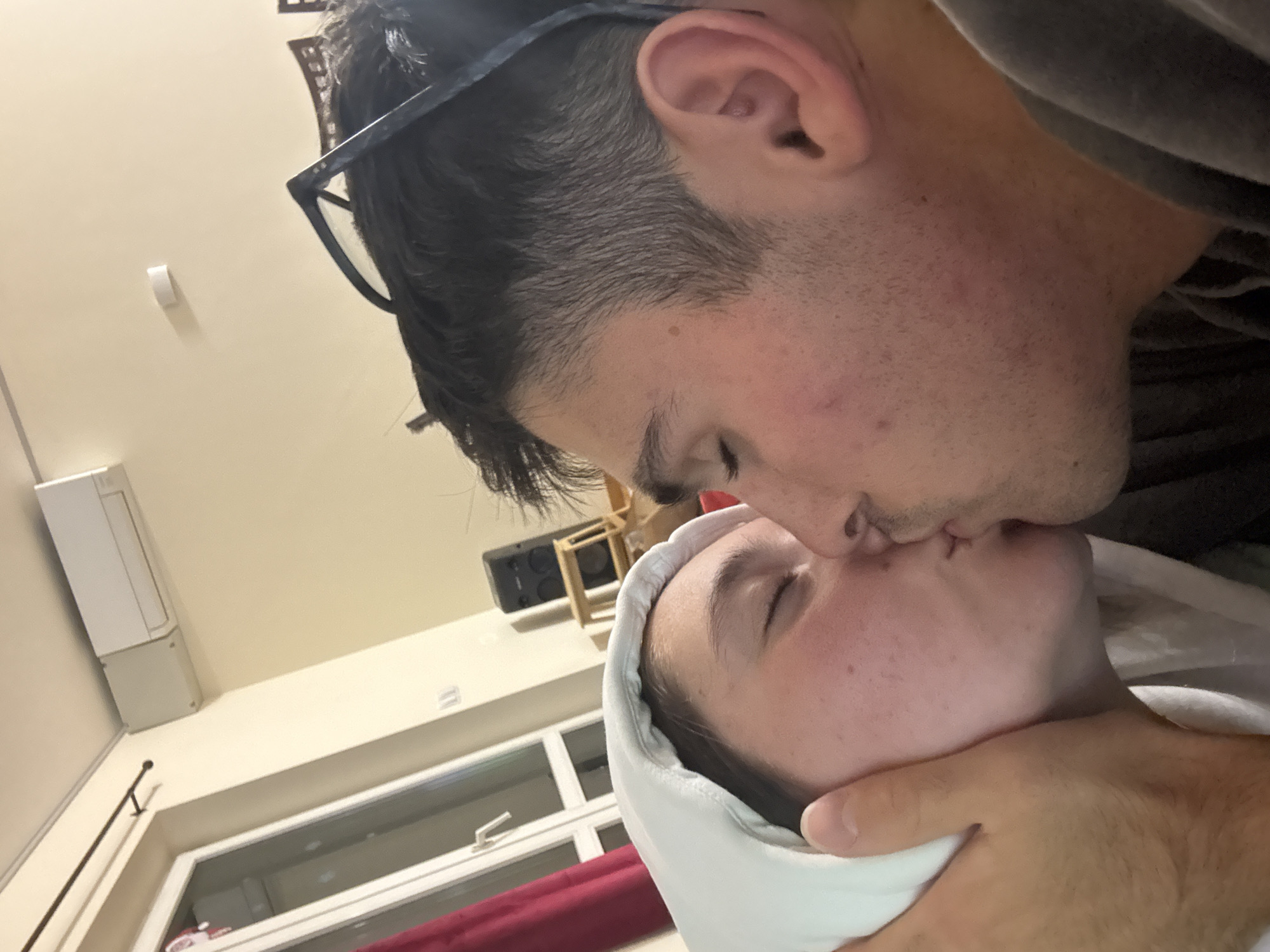 A close-up, indoor, eye-level, medium shot captures a couple kissing. The man, on the right, is leaning in towards the woman, on the left, who is wearing a white hooded sweatshirt. His hand is visible on her cheek, supporting her face. The background includes an air conditioning unit mounted on a light-colored wall, a window with red curtains, and a speaker on a wooden stand. The lighting is dim. This scene is not discernable as being in Sofia, Bulgaria.