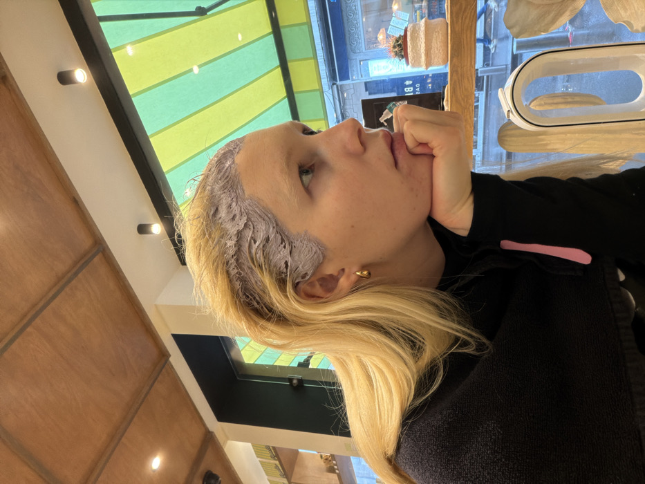 A fair-skinned individual with long blonde hair is depicted in profile, facing right, seated indoors. A light purple/grey chemical product is visibly applied to the roots and upper sections of their hair, indicating a hair treatment process. The individual's chin rests on their right hand, and they wear a black long-sleeved garment with a pink stripe on the right sleeve, along with a dark protective towel draped over their shoulders. A small, golden, teardrop-shaped earring is visible on the right ear.
The interior environment features a wooden paneled ceiling, a white ceiling section with black cylindrical spotlights, and a dark vertical partition. A large window provides a view to the exterior. Through the window, a prominent yellow and green striped awning is visible. An outdoor street scene is blurred, but includes reflections of interior elements such as a small potted cactus with red flowers in a white textured pot, and a white U-shaped device (possibly an air purifier) on an interior window ledge. Distant blurry figures and a partially visible sign containing "BI" and "18" suggest a commercial street. The street surface appears wet. The distinctive striped awning and general urban setting are characteristic of Paris, France.