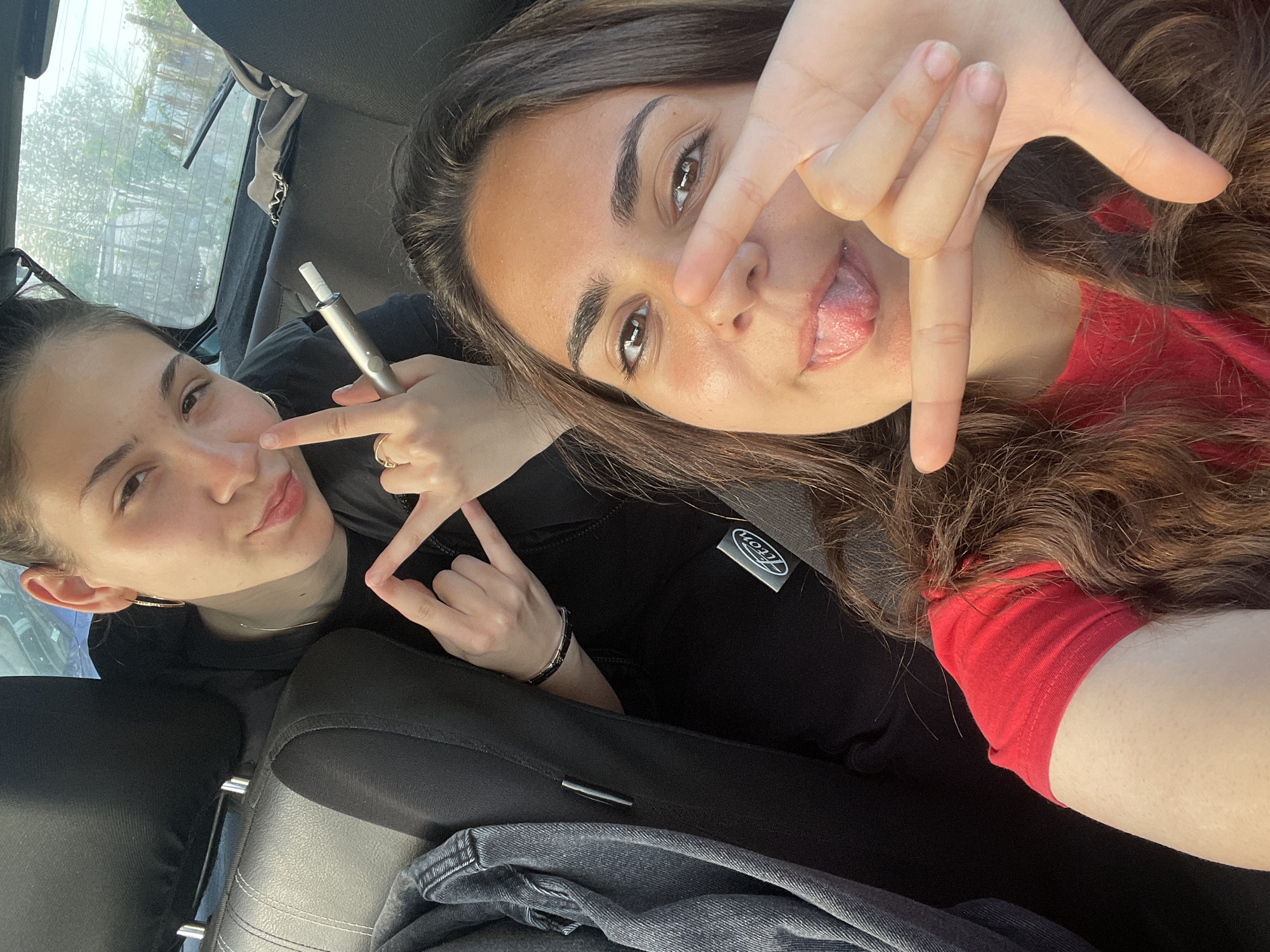 Two young women are posing for a selfie inside a car. The woman on the right is sticking her tongue out and making a hand gesture. The woman on the left is holding a metallic electronic cigarette and pointing to it with her other hand, also making a hand gesture. The car is stationary. The location appears to be a city based on the background seen through the car window.