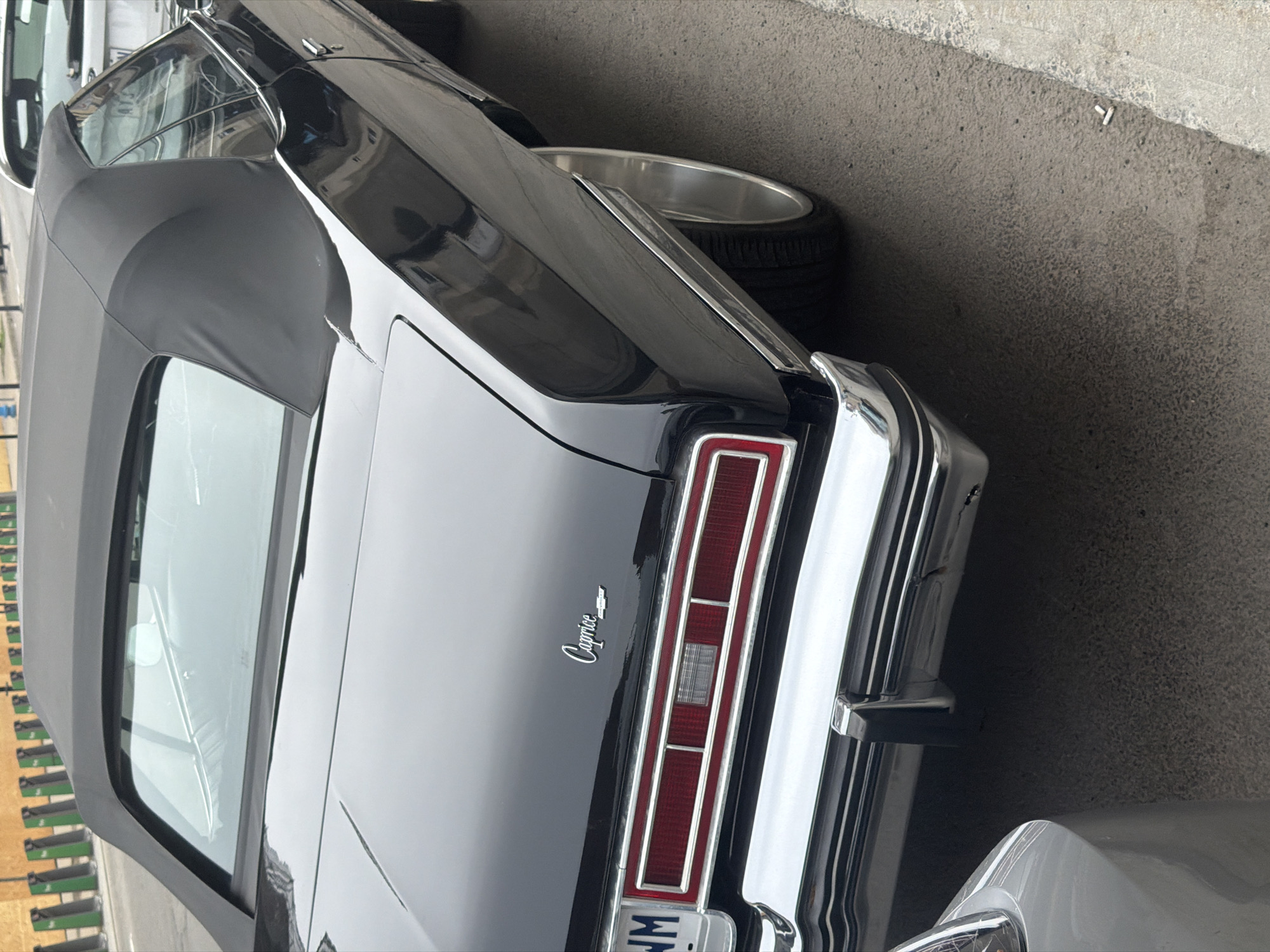 The image depicts the rear of a black Chevrolet Caprice convertible. The car is parked on an asphalt surface, with the rear taillight and chrome bumper visible. The license plate is partially obscured but appears to be a North American license plate. The background shows some green structures, possibly carts or storage units. The location is not definitively identifiable as Montreal based on the provided image alone. Further visual cues such as signage or architectural styles would be needed to confirm the location.