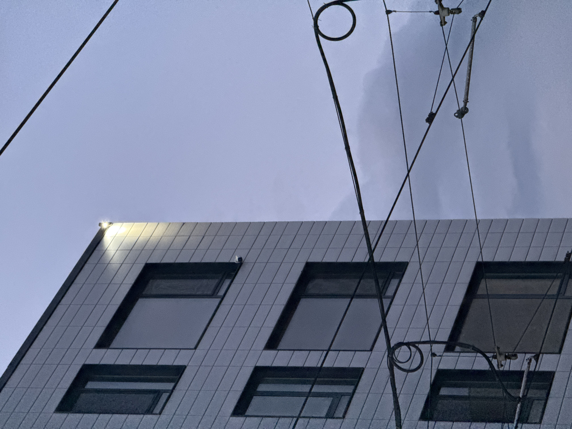 Captured from a low angle, the image features the angular upper section of a modern building. The building's facade is composed of a grid-like pattern of tiles, with rectangular windows recessed into the structure. A security camera is visible near the top left. A bright light shines from the top corner of the building. Overhead, a complex network of power lines and electrical infrastructure intersects the scene. The sky is overcast, suggesting either early morning or late evening. Sofia, Bulgaria, is not discernible from the image.