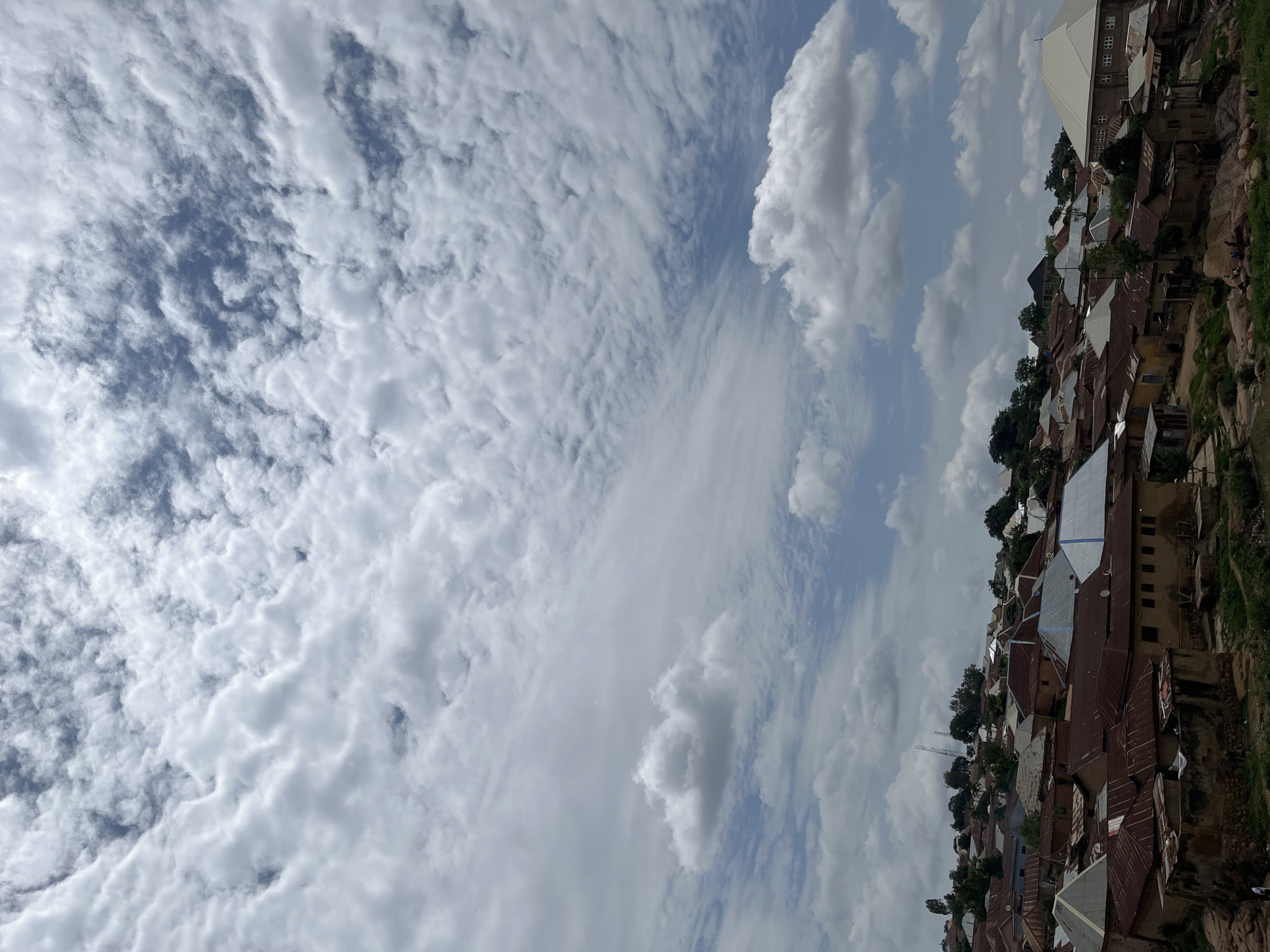 The image captures a residential area in Jos, Nigeria, under a partly cloudy sky. The upper two-thirds of the frame are dominated by a complex cloudscape: the upper portion features dense, textured, high-altitude clouds resembling cirrocumulus, partially obscuring the blue sky, while the lower sky displays scattered cumulus clouds against a clearer blue backdrop.
The lower third of the image depicts a densely packed hillside settlement. Numerous residential structures are built into the undulating terrain, characterized by a mix of architectural styles. Most houses feature corrugated metal roofs, exhibiting varying degrees of rust (reddish-brown) and some painted silver or white. The walls are typically light brown or beige. In the mid-ground, a prominent building with a large, light-colored pyramidal roof is visible on the right. Towards the upper left, on a higher elevation, several tall, slender structures, possibly communication towers, rise above the tree line. The foreground reveals rocky outcrops and sparse vegetation interspersed among the dwellings. A few indistinct human figures are visible on the rocky ground in the lower-left corner, suggesting limited activity.