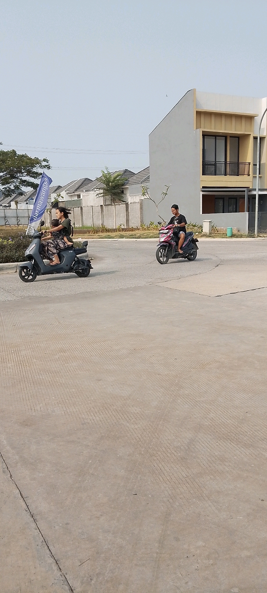 In Satriamekar, Indonesia, two individuals are riding scooters on a paved road. On the left, a woman is driving a dark gray scooter, with a child seated behind her. The woman is wearing a dark floral-patterned dress and sandals. The child, positioned behind her on the scooter, is wearing a green t-shirt and dark shorts, with what appears to be a backpack. A blue and white flag is attached to the front of their scooter. On the right, a man is riding a pink and black scooter. He is wearing a black t-shirt and dark shorts. The road is bordered by a sidewalk and a low concrete wall with some vegetation. Behind the wall are houses with gray roofs and light-colored facades, and a modern multi-story building with cream and yellow accents. The sky is clear and light blue, suggesting a daytime scene. The foreground is a concrete surface with a textured pattern of parallel lines.
