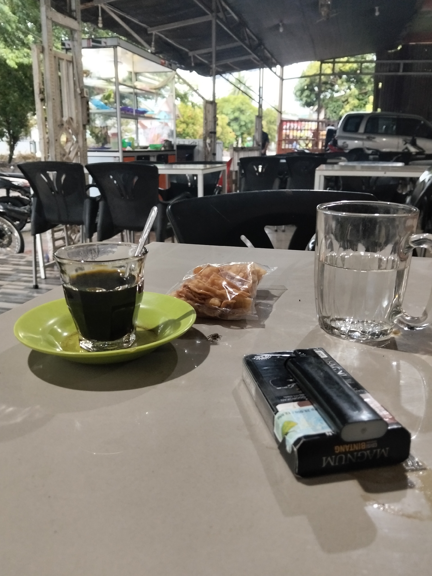The image captures a casual, semi-outdoor cafe or eating area, likely in Tanjonghajimuda, Indonesia. In the foreground, a light-colored table holds several items. A glass of dark coffee, with a spoon resting inside, sits on a green saucer. Beside it, a small clear plastic bag contains crispy, light-brown snacks, possibly kerupuk. A small, dark insect is visible on the table near the snacks. To the right, a clear glass mug is filled with water. Further right, a black pack of "MAGNUM BINTANG" cigarettes, with partially visible Indonesian government health warning text on its side, has a black lighter resting on top.
The background reveals the setting: black chairs with metal frames are arranged around white tables. Further back, under an open-sided structure with a metal frame roof, a glass display cabinet, possibly for food, is visible. Beyond this, green foliage from trees and parts of parked vehicles, including a silver SUV and motorcycles, can be seen. The lighting suggests it is daytime, possibly late afternoon, creating soft shadows. The overall scene depicts a quiet, unhurried moment at a local establishment.