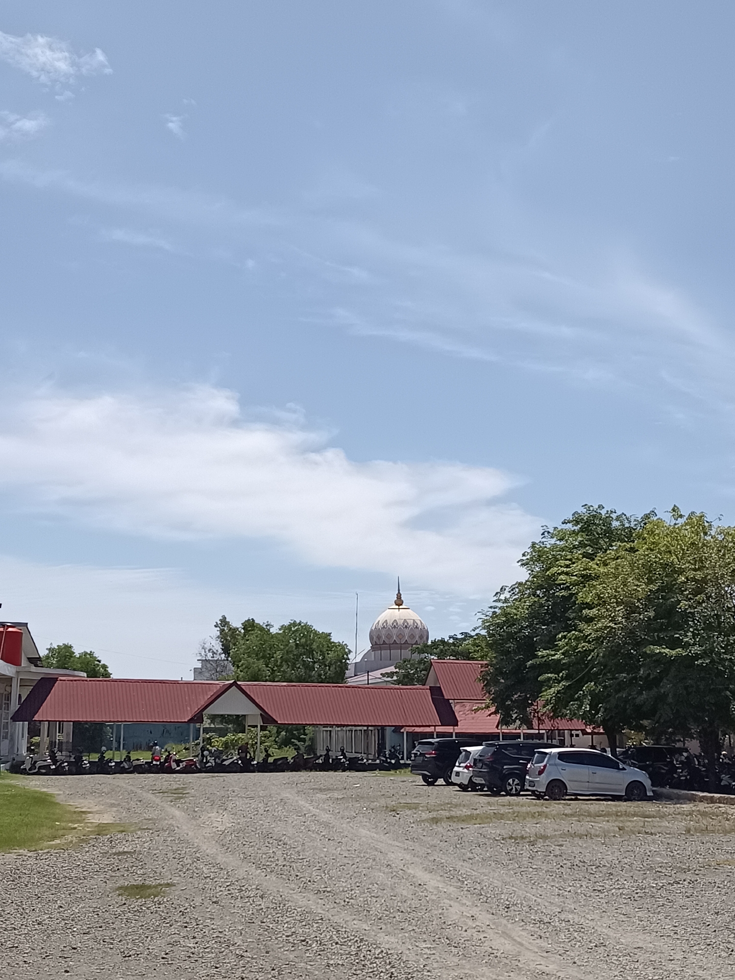 The image depicts a parking area with a gravel surface in front of a row of buildings. The buildings have red, sloped roofs and white walls. There are several motorcycles parked in front of the buildings, and a few cars are parked further to the right. Lush green trees are visible behind the buildings, and a dome-shaped structure, possibly part of a mosque, is visible in the distance. The sky is blue with scattered white clouds. The scene appears to be in Mesjid Reuntoh, Indonesia, given the presence of a mosque-like structure and the local architectural style.