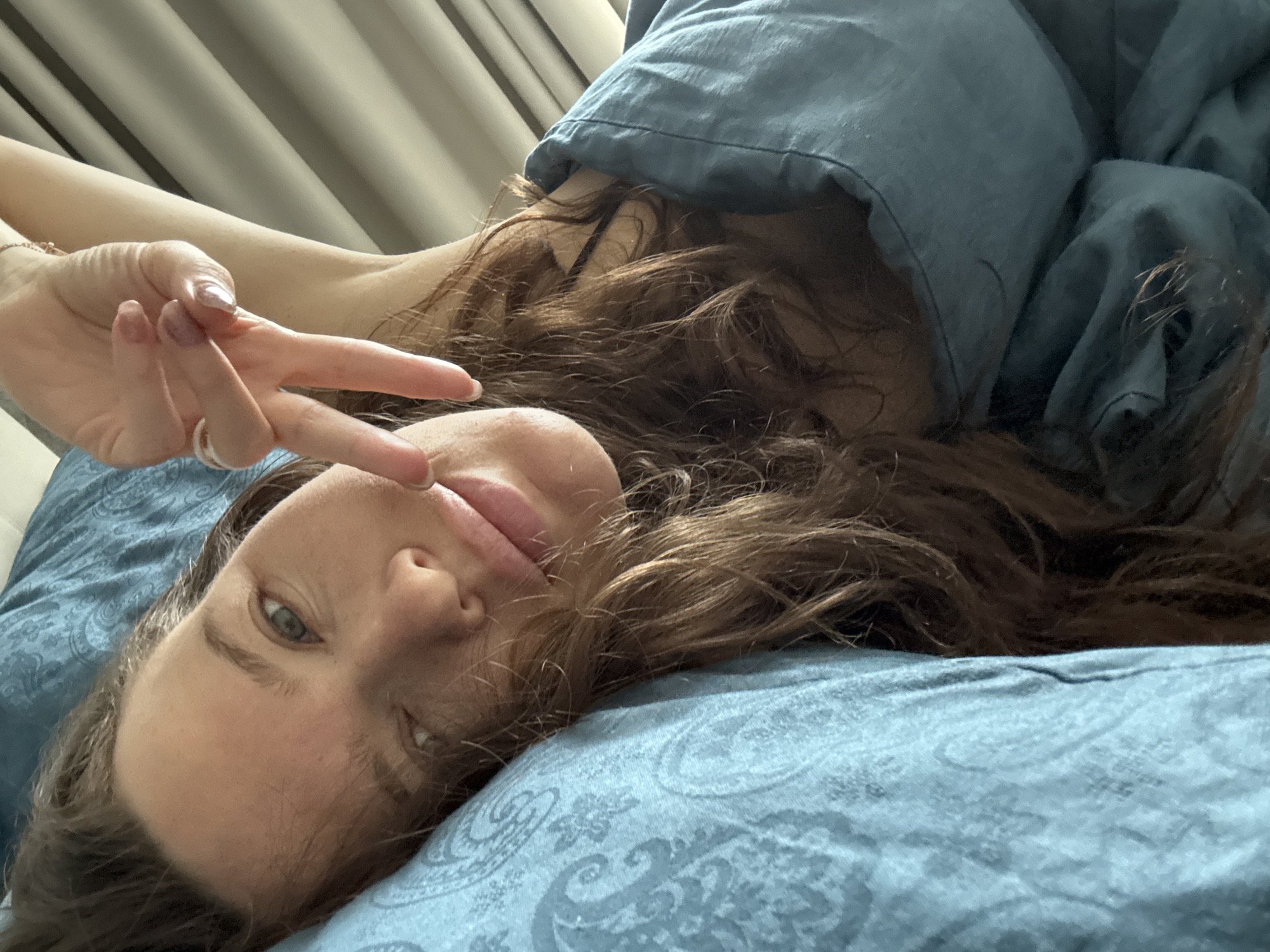 A woman lies on a blue patterned bed, partially covered by a blue duvet. She is holding up her hand in a peace sign near her face. Her hair is long and brown. The background includes a window with closed curtains. It is impossible to determine if the location is in Khimki, Russia, or any other specific city.
