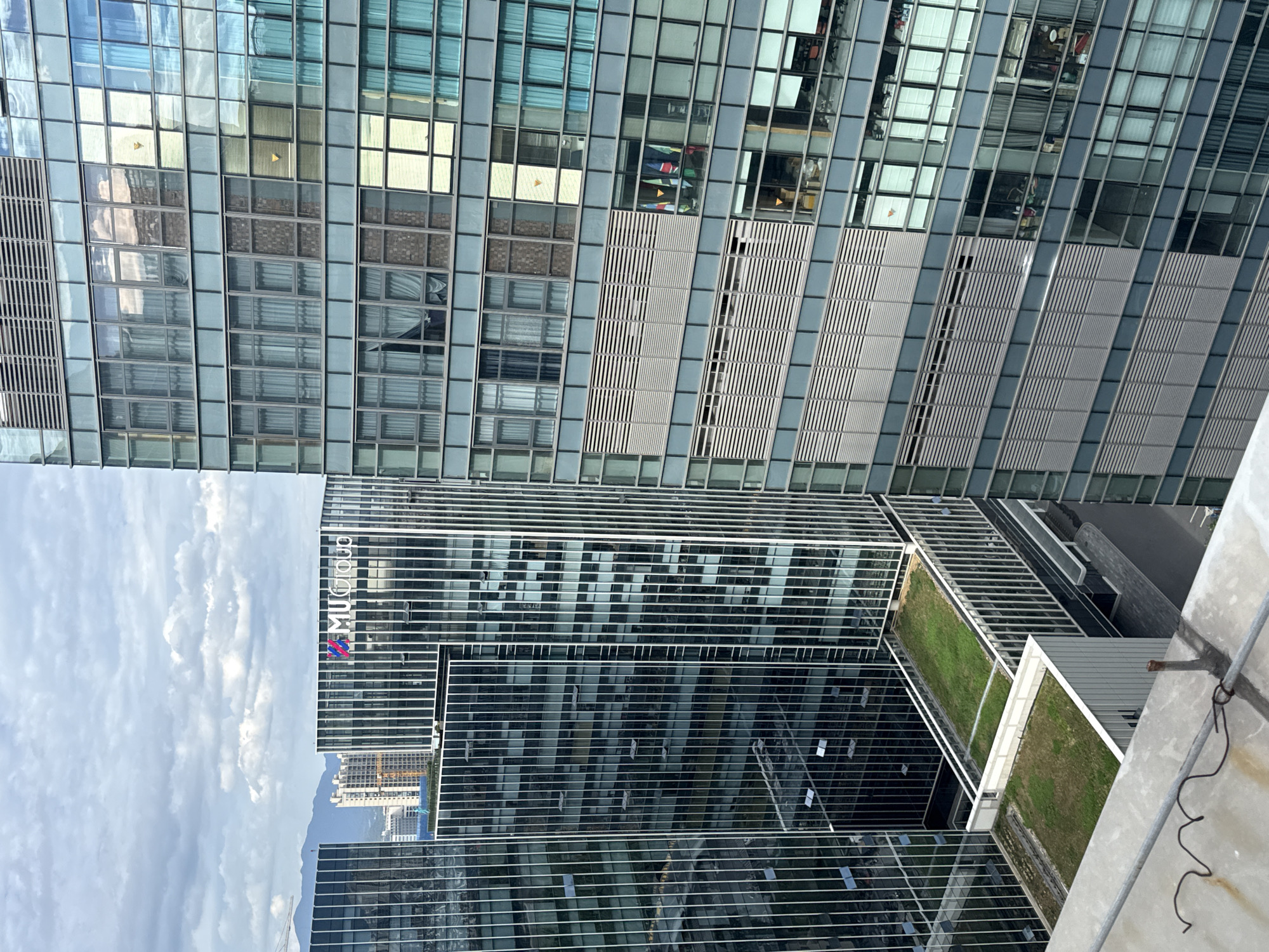 The image shows a modern cityscape in Yiwu, China, dominated by tall, glass-facade office buildings. In the foreground, a concrete ledge with some exposed wires and a rusted metal post is visible. Beyond this, a building with a green roof is apparent, set against a backdrop of several high-rise structures. One building in the middle ground displays the "MUCGroup" logo. The windows of the buildings reflect the sky, clouds, and surrounding structures. Some windows show interior details such as blinds, curtains, and office equipment. Several flags are visible hanging from a window in one of the buildings. In the distance, mountains can be seen under a cloudy sky, along with other buildings and construction cranes, indicating ongoing development.