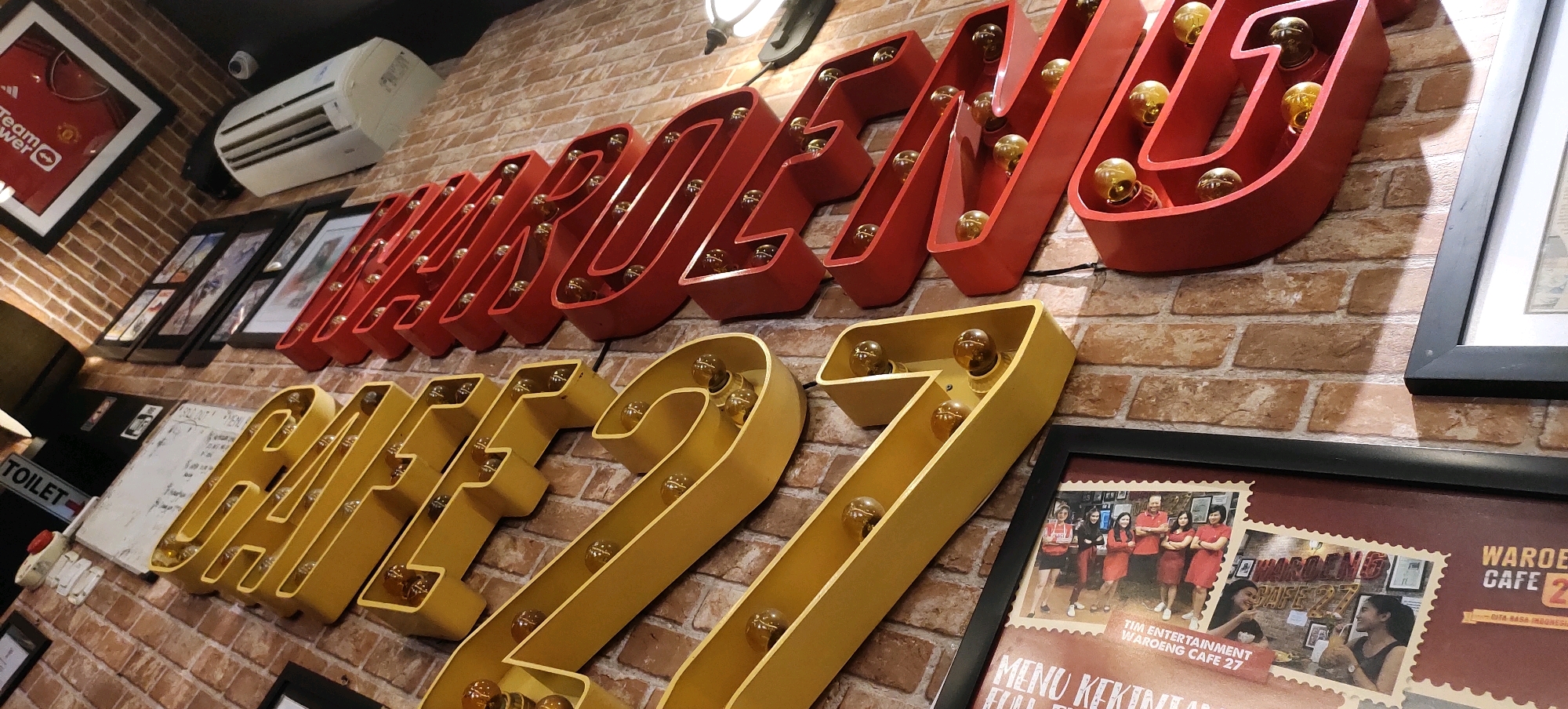 The image depicts the interior of a cafe, identified by signage as "WAROENG CAFE 27". The walls are adorned with decorative elements, including large, illuminated red letters that spell out a word, possibly part of the cafe's name or a slogan, and large yellow numbers "CAFE 27" with embedded lights. Several framed pictures and posters are also visible on the brick wall. One poster displays a group of people in red outfits, and another shows the cafe's name. A white air conditioning unit is mounted on the wall. The cafe appears to be located in Nganguk, Indonesia, as indicated by the poster text. The scene is captured at a slightly angled perspective, emphasizing the signage and the brick texture of the wall.
