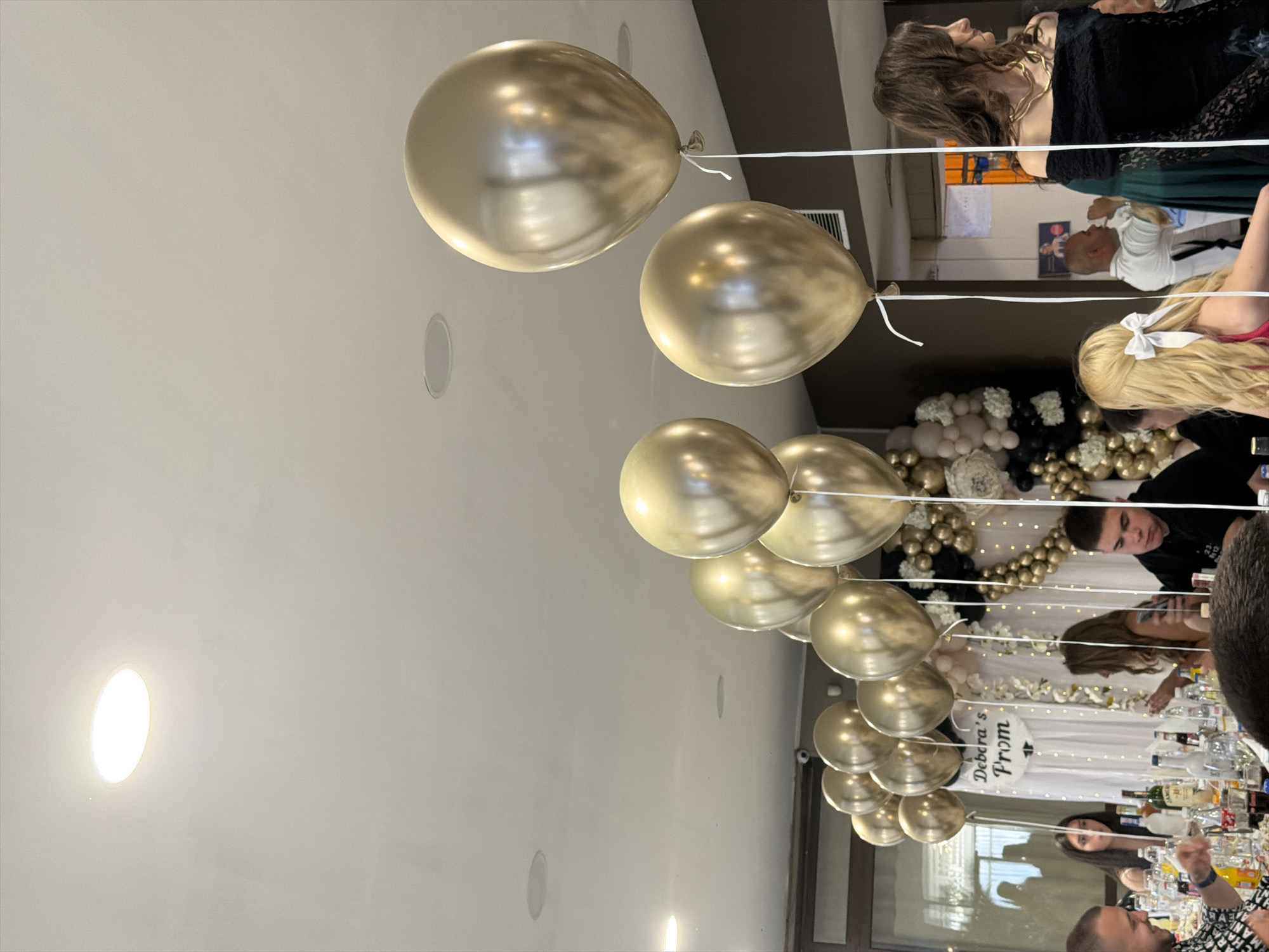 The image shows an event, possibly a prom, with gold balloons floating above tables. There are people sitting at the tables, including individuals with blonde hair and others dressed in black. In the background, there is a decorated area with more balloons and a sign that reads "Debora's Prom." Bottles and glasses are visible on the tables, suggesting a celebratory meal or reception. The location is likely Kostinbrod, Bulgaria, but there is no specific landmark or detail that confirms it.