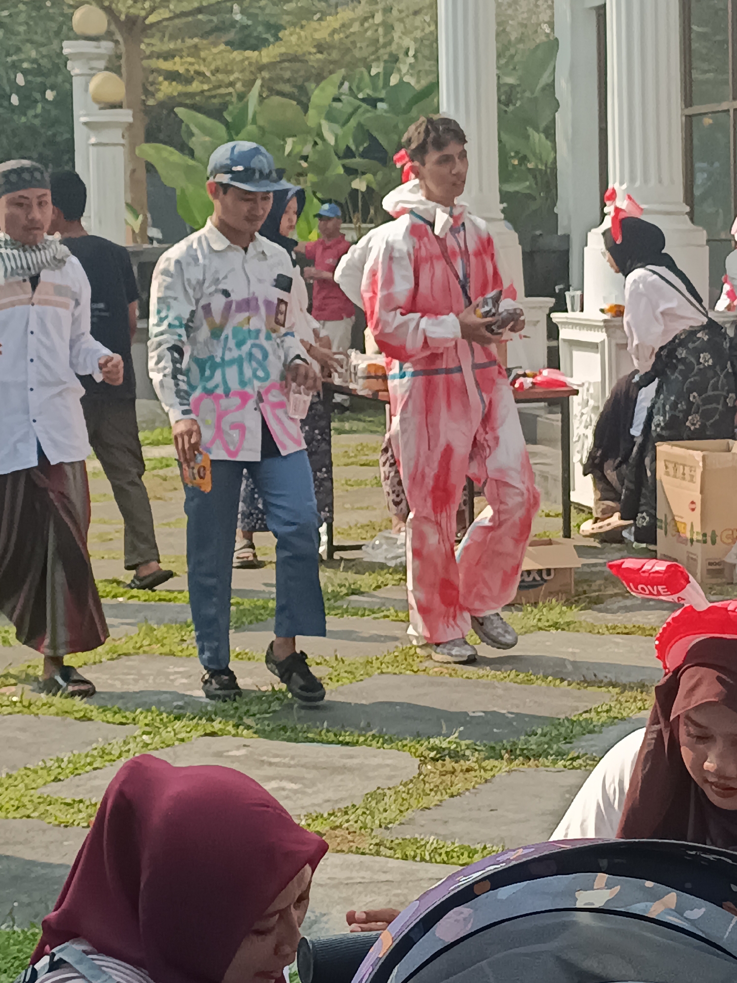 The image depicts an outdoor gathering, likely a festival or event, in Sleman, Indonesia. In the foreground, a person wearing a red and white tie-dye jumpsuit with red bows in their hair holds several small items. To their left, a person in a white shirt adorned with graffiti and a blue cap is visible. Further left, another individual in a white shirt and a sarong stands. In the background, other people, white pillars, lush green foliage, and tables with items are present. A red inflatable object with the word "LOVE" is also visible in the lower right portion of the image.