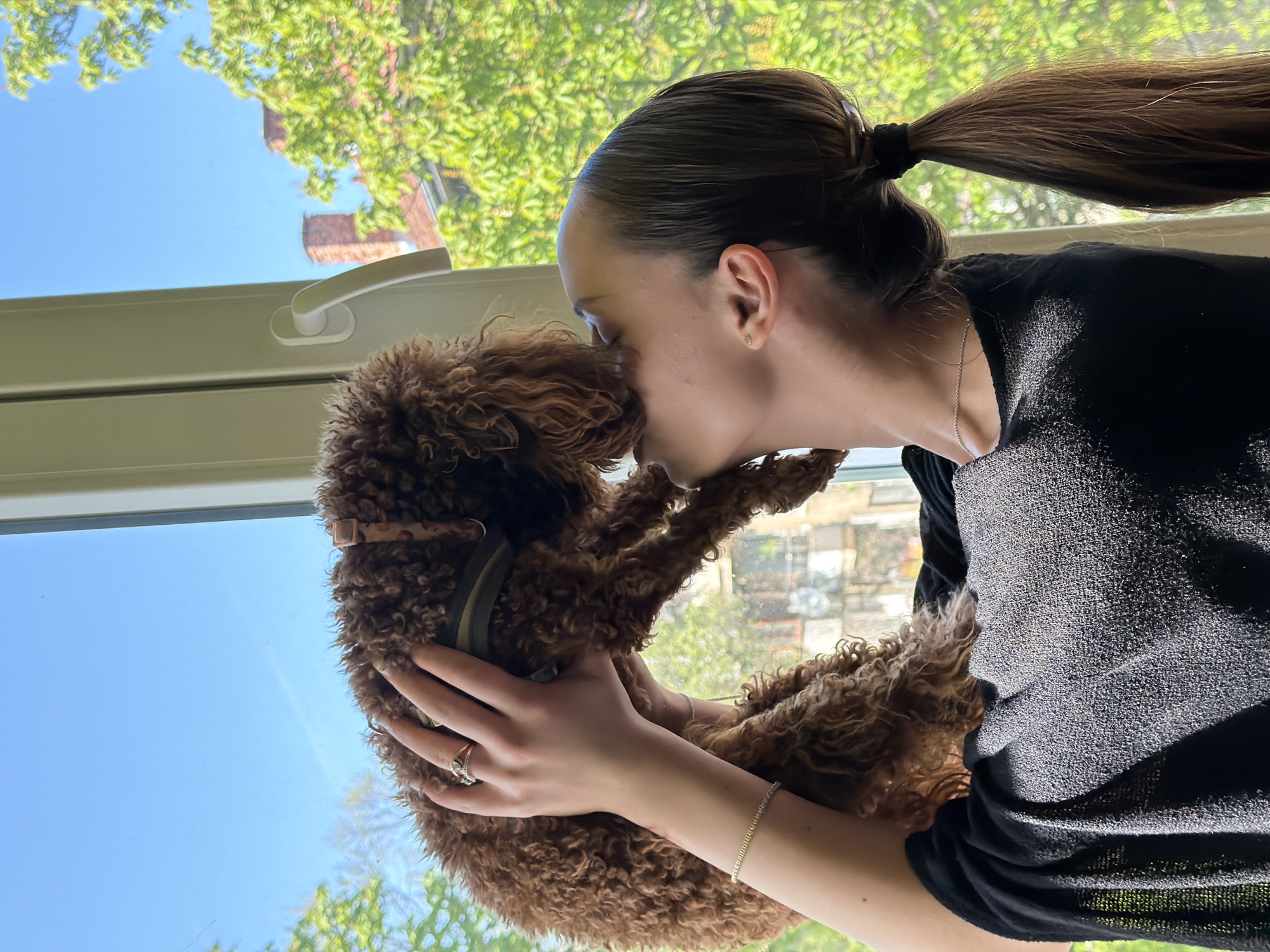 A young woman is holding and kissing a brown poodle in front of a window. The woman has dark hair pulled back in a ponytail and is wearing a dark-colored long-sleeved shirt, a necklace, a bracelet, and a ring. The dog is wearing a collar. Through the window, a blue sky and green trees are visible, along with the brick building, a characteristic feature of residential architecture in Sofia, Bulgaria. The window appears to be in a residential building. The specific location in Sofia cannot be determined from the image.