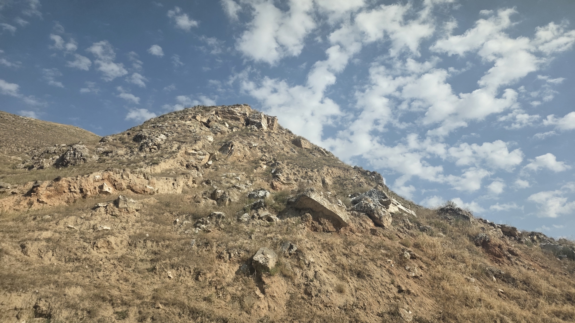 A low-angle, wide shot captures a rocky, arid mountainside under a bright blue sky with scattered white clouds. The mountain dominates the frame, its rough terrain covered in dry, brown vegetation and exposed rock formations. The soil is a dusty brown and reddish-brown, indicating a dry climate. The sky is clear and bright, with fluffy cumulus clouds dispersed across it, suggesting a sunny day. The scene is devoid of human presence or any man-made objects, presenting a natural, undeveloped landscape. The image appears to be taken on a clear day, likely during daylight hours. The location is the mountainous outskirts of Taldyk, Kyrgyzstan.
