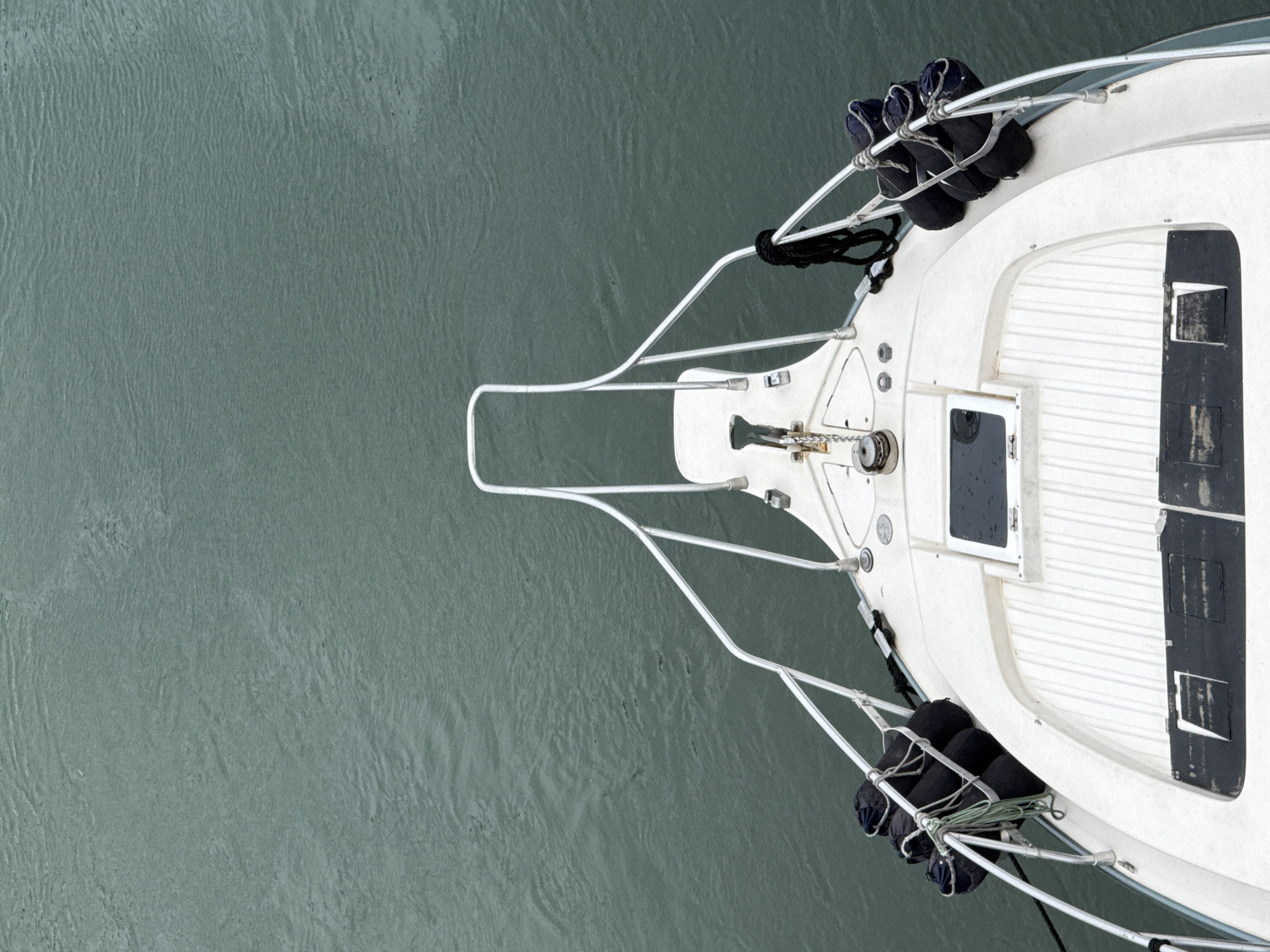 An aerial, full shot shows the bow of a white motorboat floating on calm, green water. The boat's bow curves upward with a white deck and metal railings. Several black fenders are attached to the railings on either side of the bow, secured with ropes. In the center of the bow, an anchor is visible, attached to a chain. The water is textured with subtle ripples. The scene is static and there are no people visible. The location is not specified in the image, and therefore cannot be provided for Cabo Frio, Brazil.