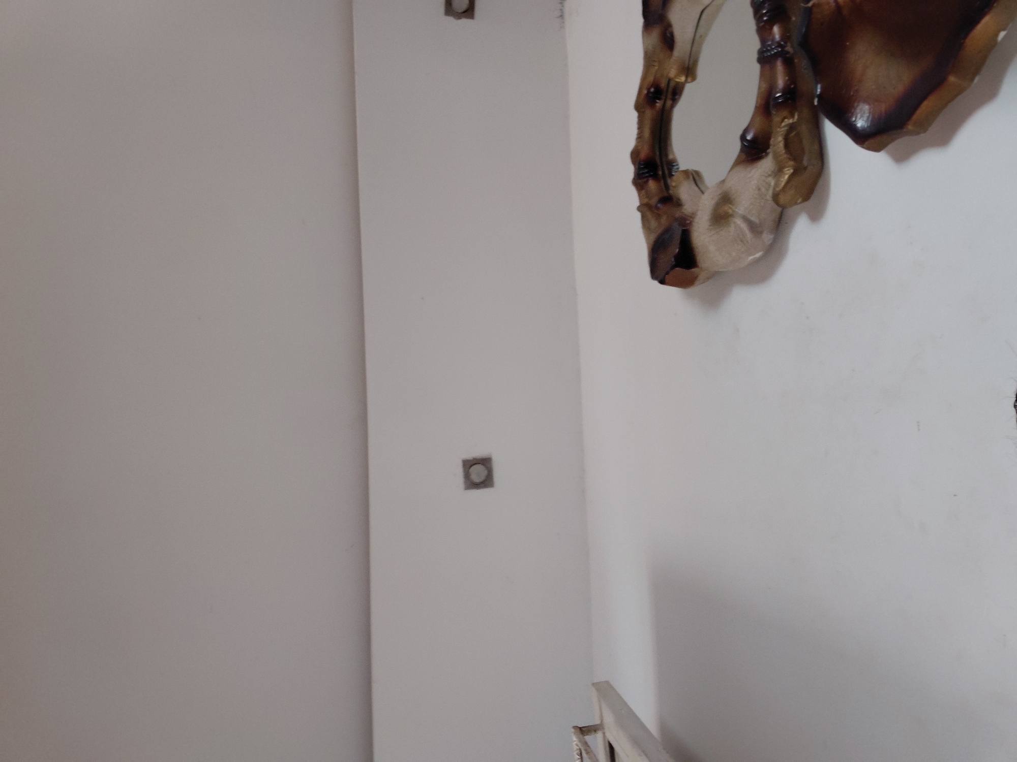 This indoor image presents a static view of a corner or wall section, likely within a building in Santa Ana, Venezuela. The primary subject is a plain white wall that takes up most of the frame, showing a minimalist aesthetic.
On the left side of the wall, two square openings are visible, positioned vertically. These appear to be electrical boxes or conduit openings, possibly for outlets or switches, with a circular element visible within them. The higher box is closer to the top edge, and the lower one is roughly in the middle of the left wall section.
To the right, a large, ornate decorative object or mirror frame is partially visible. It features an organic, irregular shape with rich textures and colors, predominantly shades of dark brown, burnt umber, and lighter beige or tan. The design suggests a rustic or handcrafted style, possibly resembling carved wood or a similar material.
At the very bottom right corner, a small section of a light-colored railing, possibly part of a staircase or balcony, can be seen, with vertical elements suggesting balusters. The lighting is even and diffused, typical of an indoor environment. There are no people or discernible activities taking place. No text is visible in the image. The exposed electrical boxes suggest the space might be unfinished or under construction.