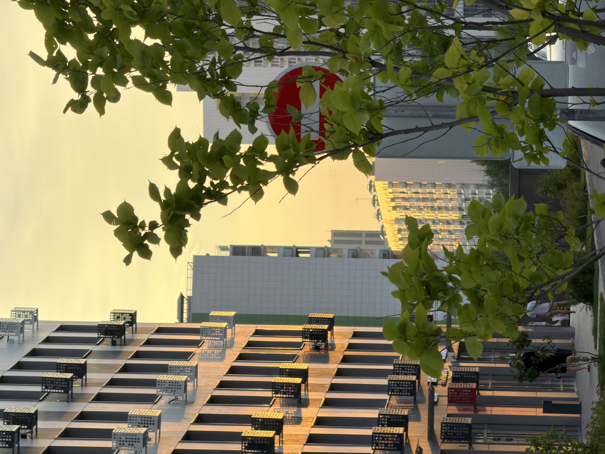 The image shows a street scene in a city with modern apartment buildings. In the foreground, there is a young tree with green leaves. To the left, a tall apartment building with balconies is visible. The building has a facade of light-colored panels and dark vertical window frames. Some balconies have dark metal railings, while others have white decorative screens. Further back, another apartment building with a white facade and rows of windows can be seen. The sky is a warm yellow, suggesting it is either sunrise or sunset. A red and white circular traffic sign is partially obscured by the tree's foliage. In the middle ground, there are two people walking on a sidewalk, one wearing a dark t-shirt and shorts, and the other in a white t-shirt. A car is partially visible in the lower right corner. The lighting indicates that the sun is low in the sky, casting long shadows and bathing the buildings in a warm glow.