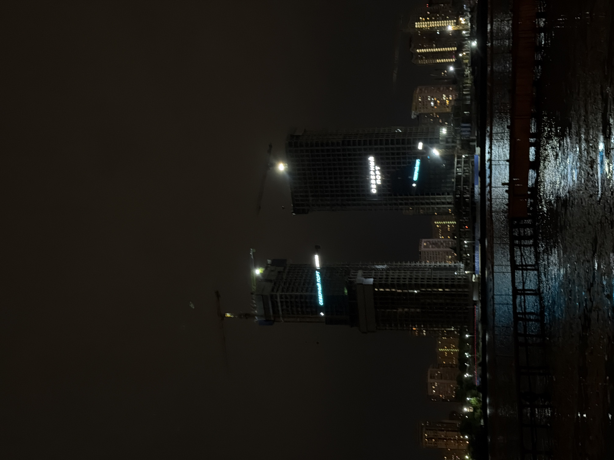 The image shows a nighttime view of the Moscow Canal with two partially constructed skyscrapers. The left skyscraper has a sign that reads "СЕВЕРНЫЙ ПОРТ" (Northern Port) in blue neon letters, and a construction crane extends from its top. The right skyscraper also has a similar sign with "СЕВЕРНЫЙ ПОРТ" in white neon letters. In the background, several other residential buildings with illuminated windows are visible, suggesting an urban environment. The water in the foreground reflects the lights from the city and the buildings, creating a shimmering effect on the surface. A wooden pier or walkway is visible in the lower part of the image, partially submerged in the water. The sky is dark and overcast, with no stars or moon visible. The scene depicts an ongoing construction project in an established urban area.