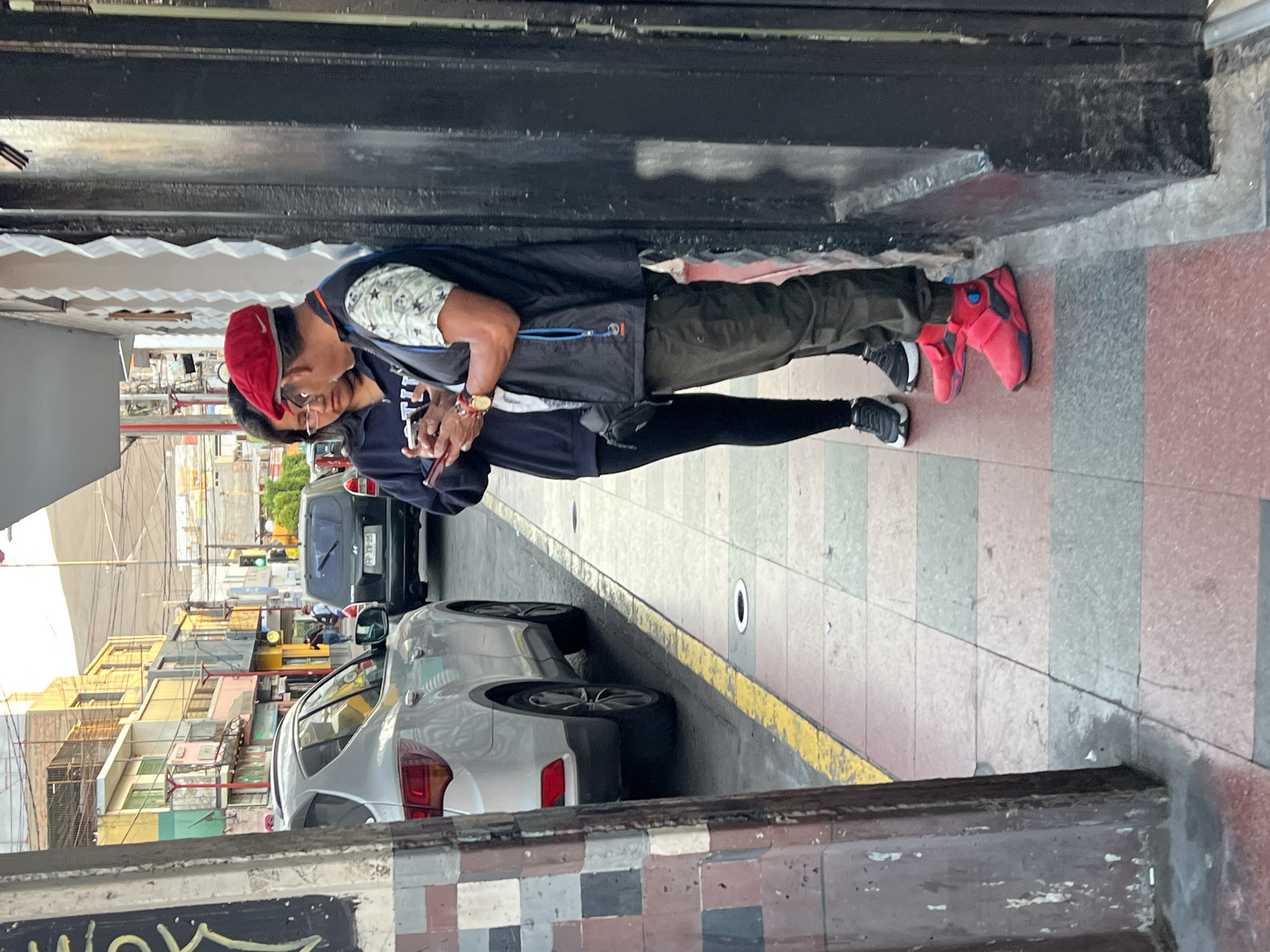 A photograph captures a street scene in Arica, Chile. Two people stand on the sidewalk next to a parked silver car. The person closest to the camera wears a red cap, glasses, a patterned shirt, dark vest, and dark pants, while looking at a mobile phone in their hands. The second person wears a dark jacket and leggings with black and white athletic shoes. Parked cars line the street, and multi-story buildings can be seen on the left side of the frame. The sidewalk has gray, red, and white vertical stripes. The photograph is taken from within or very close to a doorway, with sections of the doorway visible on the top and bottom.