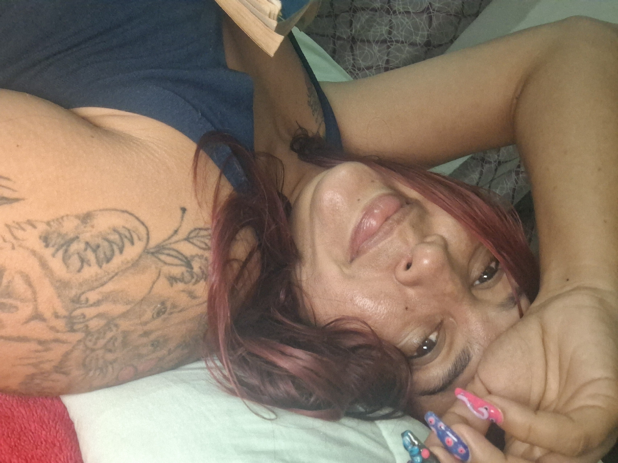 A woman with red-streaked hair is lying down on what appears to be a bed with a patterned sheet. She has an extensive tattoo on her left arm and is wearing a blue top. Her hand is near her face, showcasing long, decorated nails with pink and blue designs. She is looking at the camera. No information in the image allows the specification of the city of Vallecito, Venezuela.