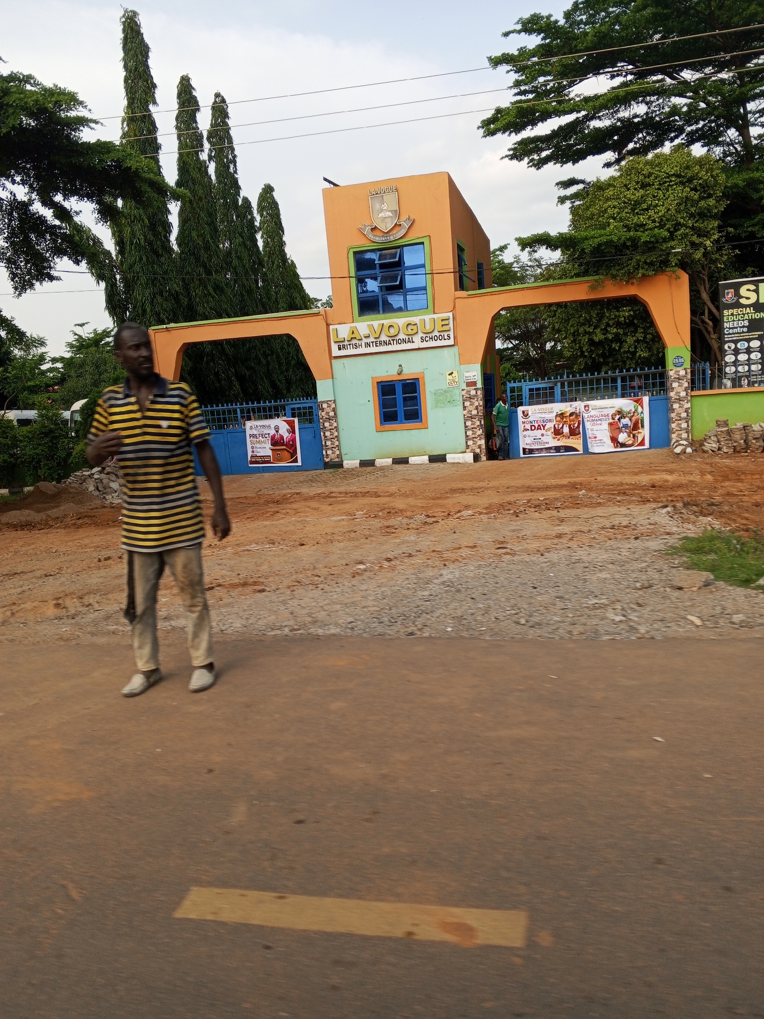 In Karu, Nigeria, the image captures the entrance to "LA VOGUE British International Schools." A man in a yellow and black striped t-shirt and khaki pants stands on a dirt road in the foreground, facing slightly away from the camera. Behind him, the school's orange and green arched gate stands prominently, with a two-story building featuring blue-framed windows visible above. Several posters are displayed on the blue gates, promoting "LA VOGUE Montessori Day" and "Language & Humanities Week." Tall, slender evergreen trees flank the school building. The ground leading up to the gate is a mixture of dirt and gravel, suggesting an unpaved area. The sky above is overcast. A partially visible sign on the right side of the gate reads "SH SPECIAL EDUCATION NEEDS Centre."