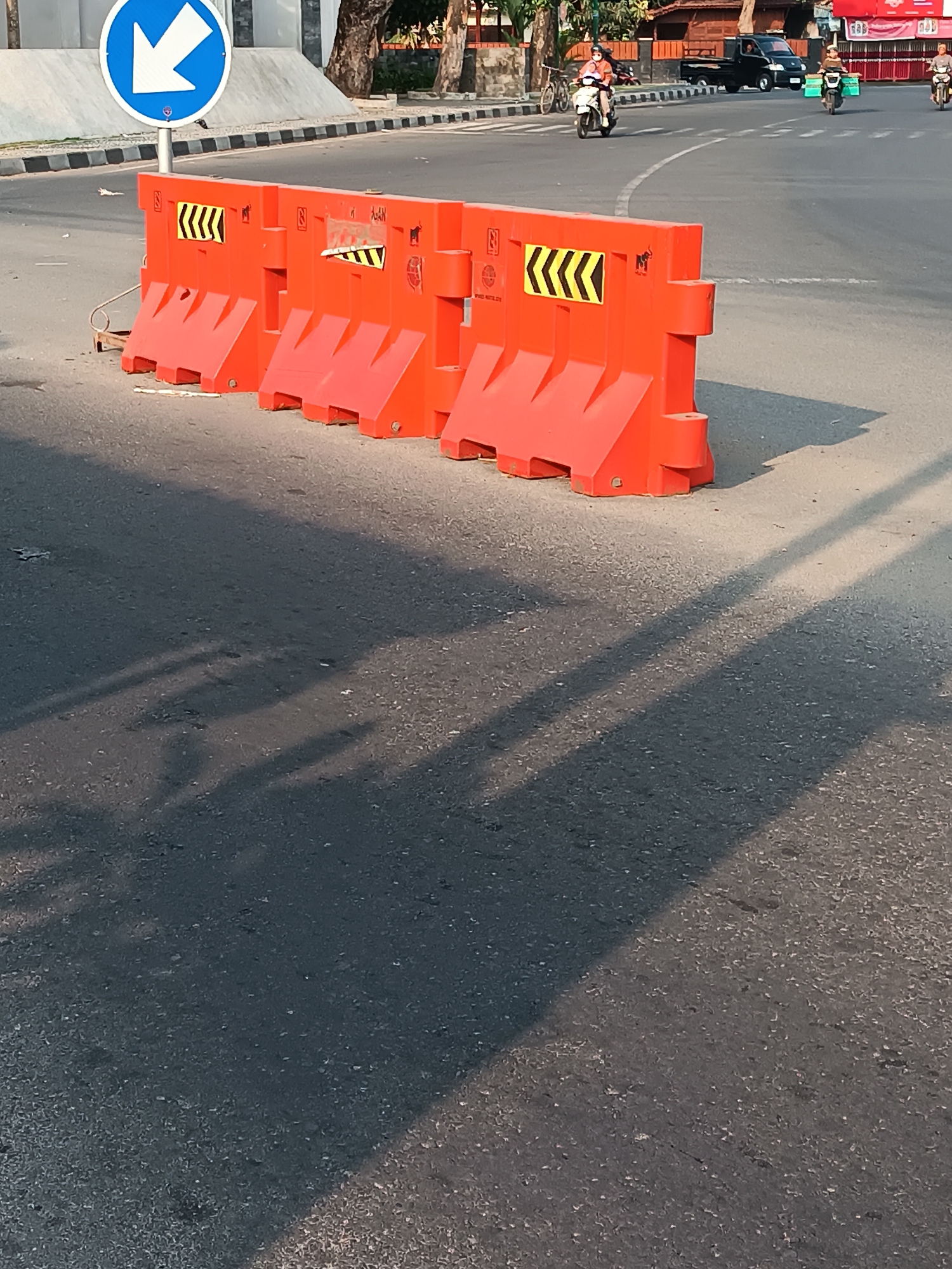 An outdoor, eye-level, medium shot captures a street scene in Imogiri, Indonesia, under bright, sunny conditions. The foreground is dominated by an asphalt road and a row of three bright orange, modular traffic barriers. These barriers, made of plastic, are interconnected and feature reflective yellow and black chevron warning stickers. One barrier on the far left has a blue circular traffic sign with a white arrow pointing left. Behind the barriers, the road curves, and several vehicles are visible. A black pickup truck is in the middle ground, and two motorbikes are traveling on the road, one in the middle ground and another further in the background. Two individuals are riding the motorbikes. In the background, there are buildings with traditional Indonesian architecture and trees. The sunlight casts long shadows across the road.