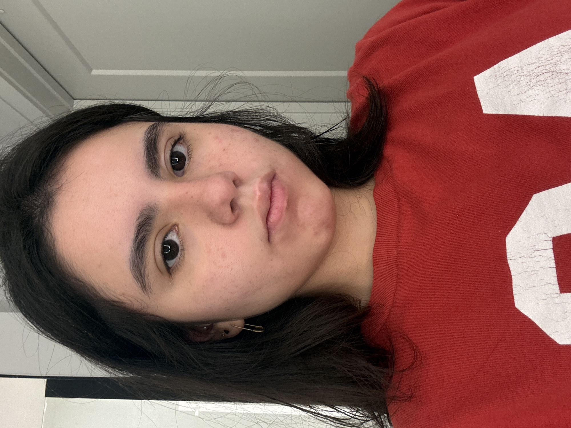 A young woman is shown in a close-up, indoor photograph. She has dark hair, brown eyes, and is wearing a red shirt with a white graphic on the right side. The photo appears to have been taken in front of a white door or wall. A safety pin is visible in her left ear. There is no evidence of location within Austin, United States of America.
