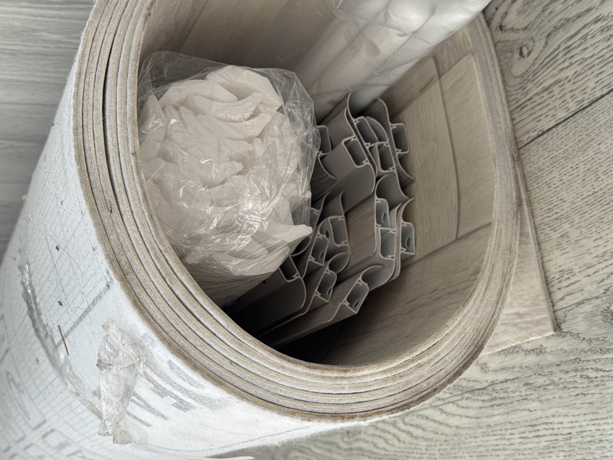 The image shows a close-up, eye-level view of a large roll of flooring material, possibly vinyl or laminate, made of multiple layers. The roll is unfurled and placed on a light-colored wooden floor with a visible grain pattern. Inside the curve of the roll, a stack of gray, U-shaped plastic trim pieces is visible, wrapped in clear plastic. There is also a crumpled white material in clear plastic packaging. The flooring material itself has a light grey pattern with darker grey lines. The background consists of a grey textured wall. The scene appears to be indoors, possibly in a storage area or a room where renovations are taking place. There are no people or animals in the image. There is no specific indication of the location being in the city of Яромінскі сельскі Савет, Belarus, other than the prompt's request.