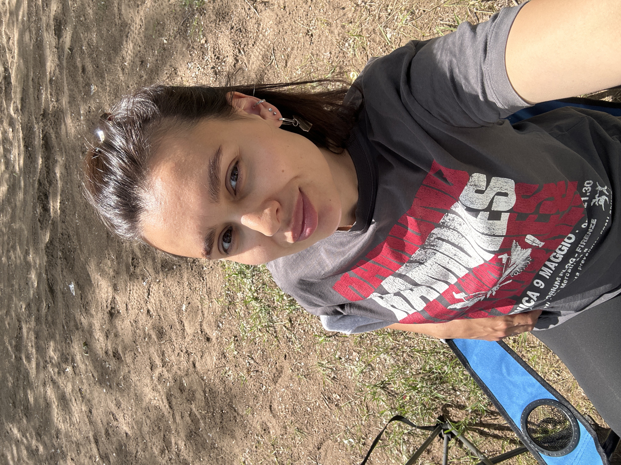 A young woman with dark hair and brown eyes is captured in a selfie outdoors. She is wearing a grey t-shirt with a red and white graphic design and black leggings. She is sitting on a blue folding chair with a cup holder. The background is composed of dirt and sparse green grass, suggesting an outdoor, possibly park-like setting. The lighting indicates it is daytime. The location is identified as Noviy, Russia.