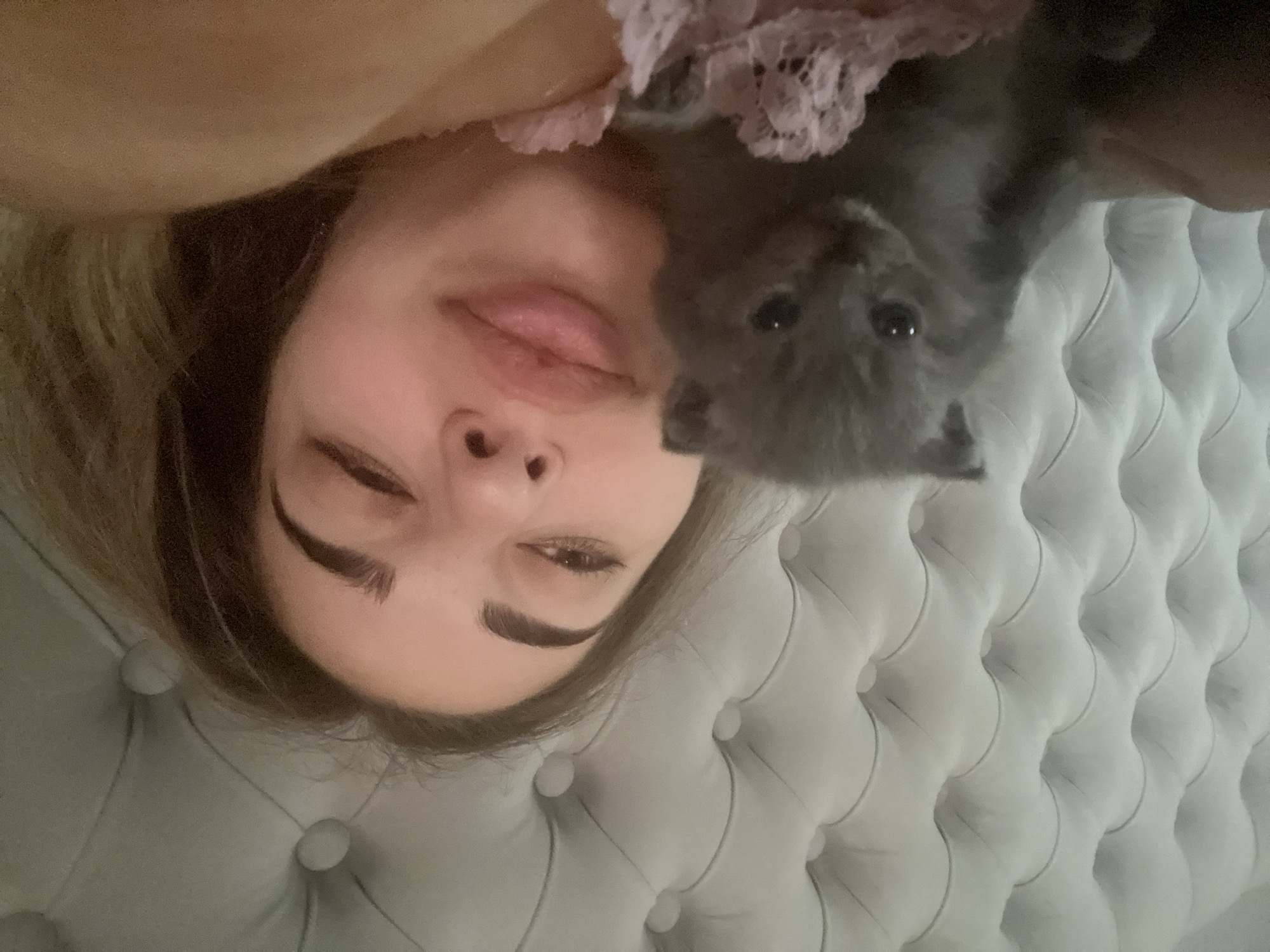A close-up, selfie-style photograph captures a young woman and a gray kitten lying in bed. The woman, positioned on the right side of the frame, is partially visible from the chest up, with her face turned towards the camera. Her eyes are open, and her lips are slightly parted. She has dark eyebrows and light brown hair. To her left, the kitten is nestled against her. It is small and gray with large, dark eyes. The background consists of a tufted, light-colored headboard, suggesting a bedroom setting. The image is taken indoors, and there is no information available about the location in Velykyi Bychkiv, Ukraine.