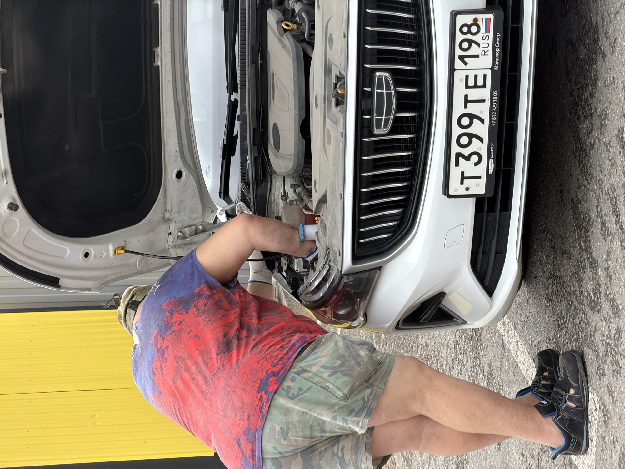 A man wearing a red and blue patterned shirt and camouflage shorts is bent over the front of a white Geely automobile, the hood of which is open. The man is facing the car, appearing to be working on the engine or the headlight area. A Russian license plate, "T399TE 198 RUS," is visible on the front of the car. The scene is set outdoors in a location identifiable as possibly being within the city of Saint Petersburg, Russia, based on the Cyrillic text on the license plate and its frame. The background includes a yellow wall or structure. The man is wearing protective gloves.