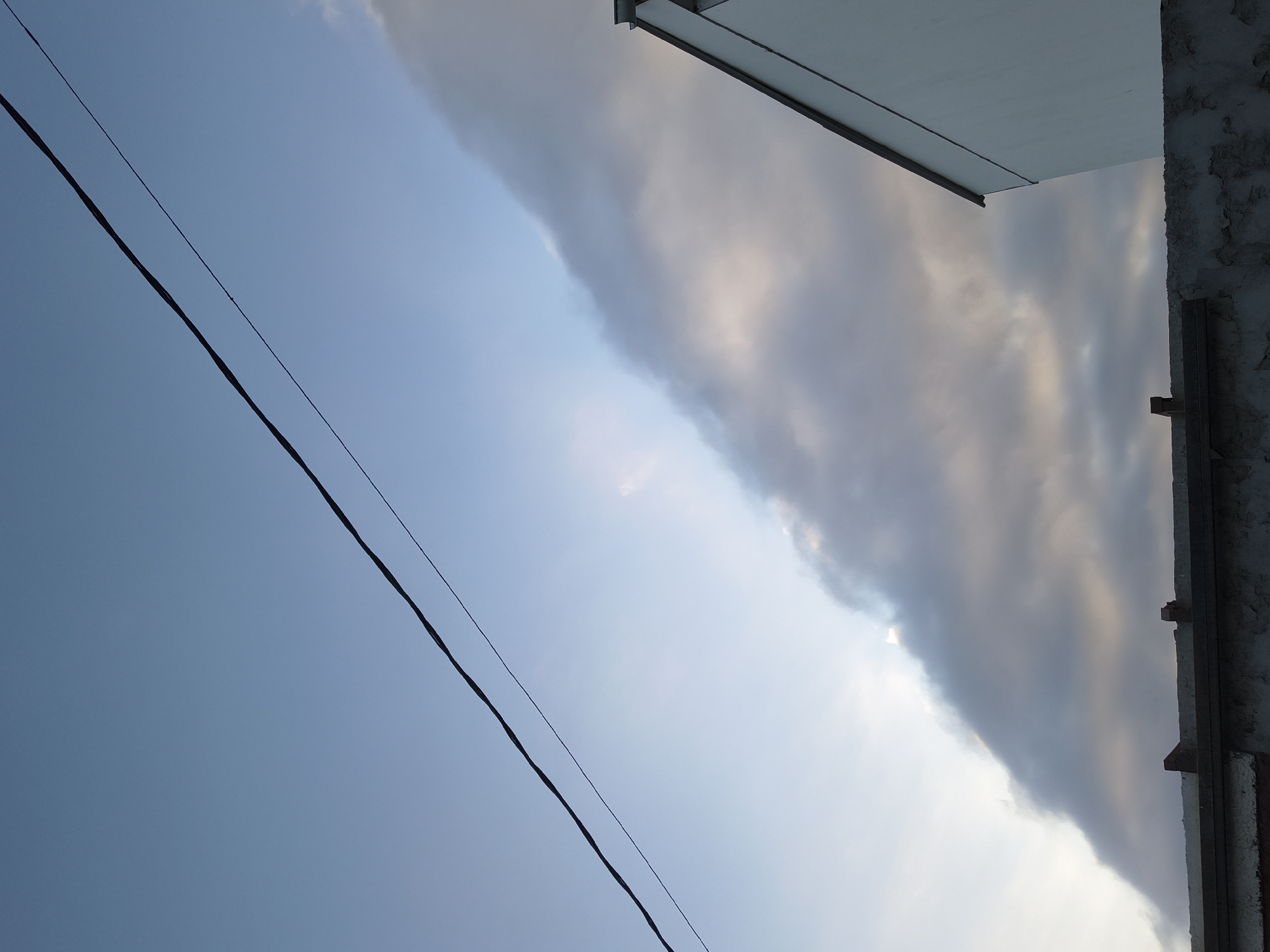 The image shows a view of the sky, an electrical wire, and the corner of a building. The sky is partially covered with a thick band of clouds, with a patch of clear blue visible on the left. An electrical wire stretches diagonally across the left side of the frame. The upper right corner features part of a building, including a white overhang and a section of textured wall with visible support beams. The location is likely Arica, Chile, given the query context. No people or actions are visible.
