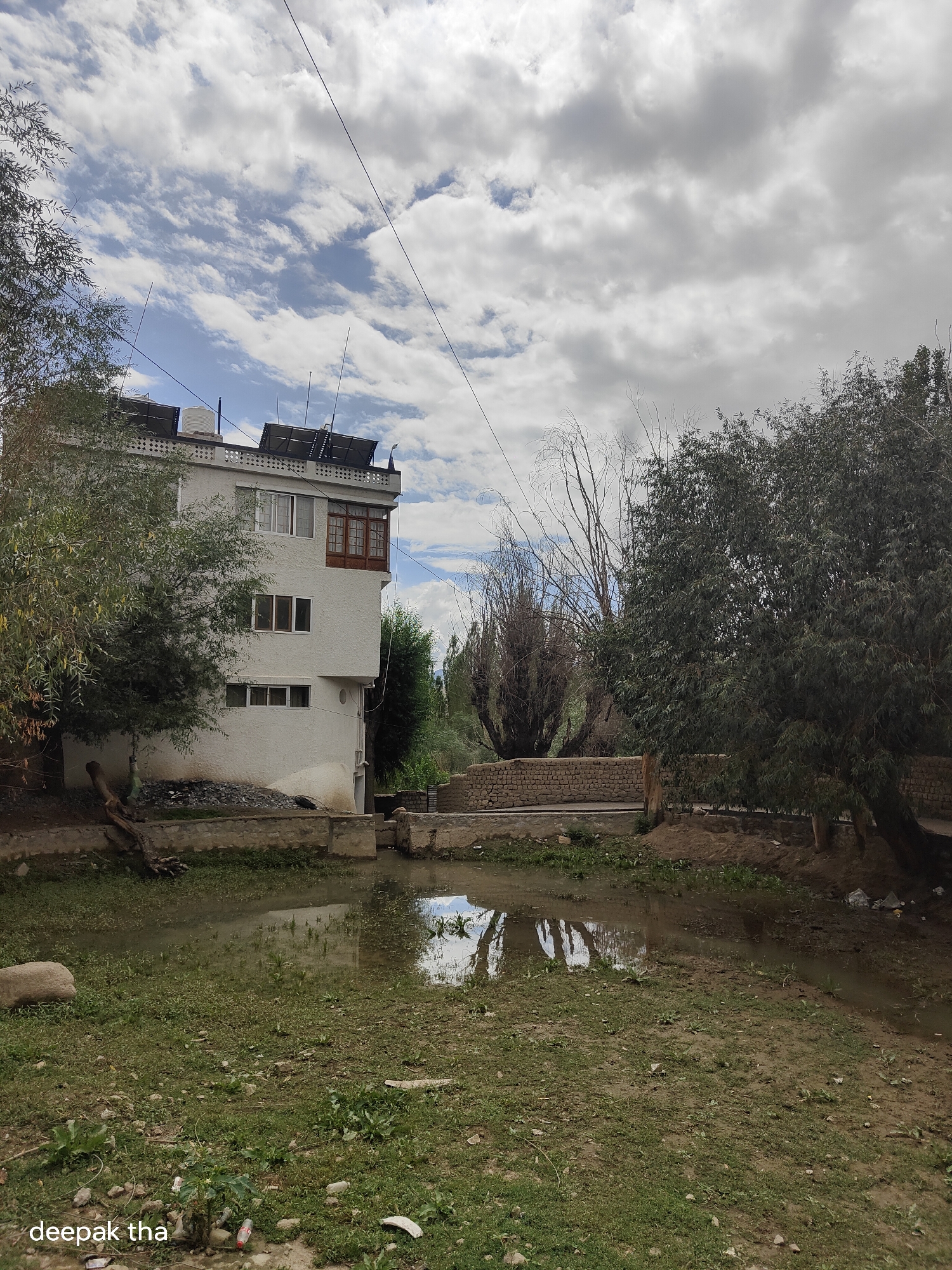 The image depicts a building and a muddy, shallow body of water in Leh, India. The building is white, with multiple windows and a balcony. Solar panels are visible on the roof, along with a water tank and antennae. A tree with green leaves is to the left of the building, and a larger, denser tree is to the right. The ground is covered in grass and dirt, with some debris scattered around. The water reflects the sky and trees. The sky is partly cloudy with a mix of blue sky and white clouds. A brick wall runs behind the body of water. The overall scene suggests a rural or semi-urban setting with signs of development and natural elements.