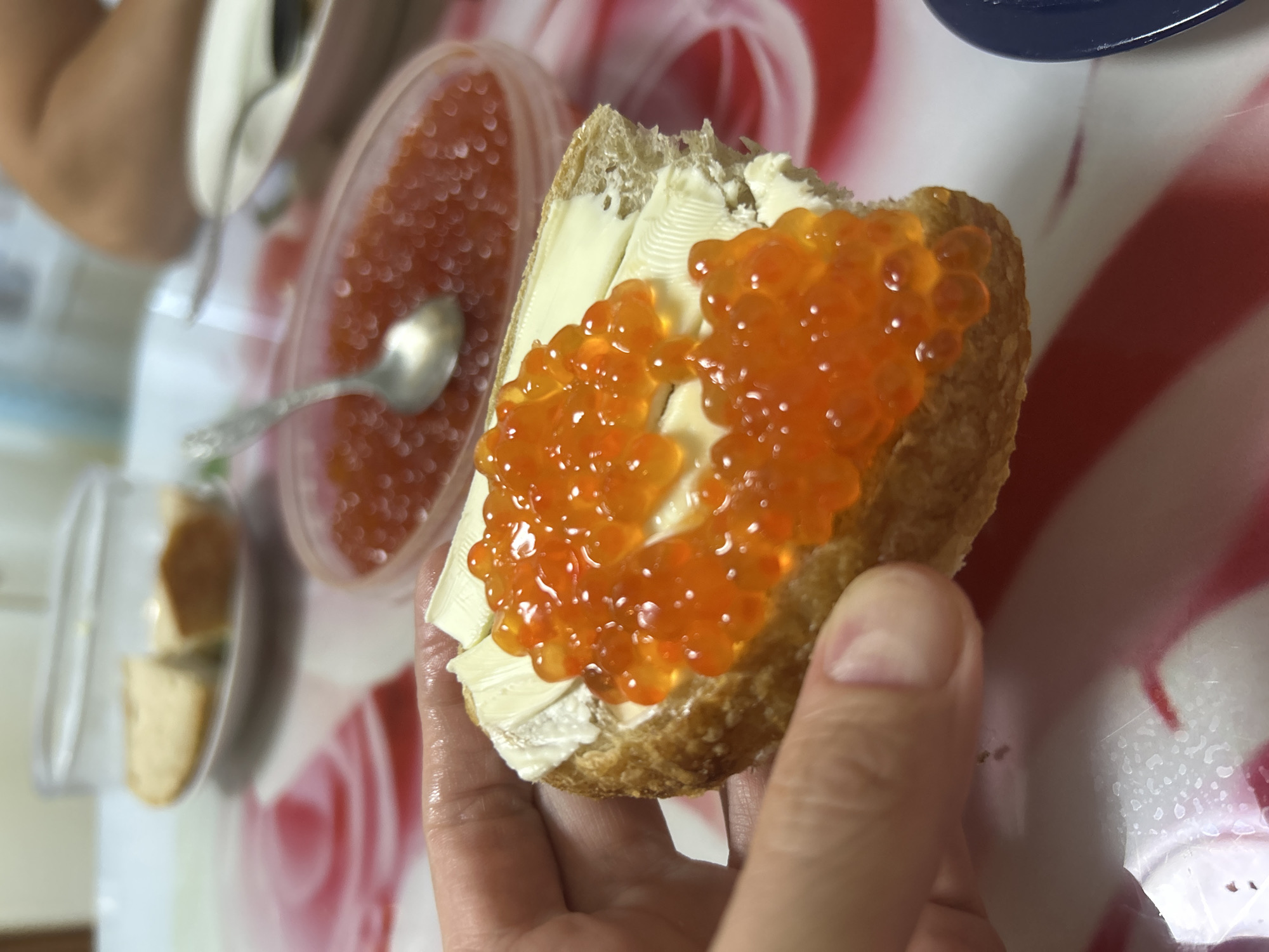 A close-up photograph captures a human hand, likely the left, holding a toasted piece of bread generously topped with a layer of pale yellow butter and bright orange-red salmon roe (caviar). A bite has been taken from the bottom right section of the bread. In the blurry background, to the upper left, a transparent dish contains additional slices of bread. To the upper right, an oval-shaped transparent container is filled with more red caviar, with a small silver spoon partially submerged within it. Further right and out of focus, parts of a white plate with a dark-handled utensil and a blurred limb are visible. The items rest on a tablecloth featuring a white base with broad, wavy red patterns. Small condensation marks or liquid droplets are present on the white areas of the tablecloth near the bottom of the frame. The setting appears to be an indoor dining environment. No specific details within the image allow for the identification of its location as Izhevsk, Russia, or any other particular city or geographic region.