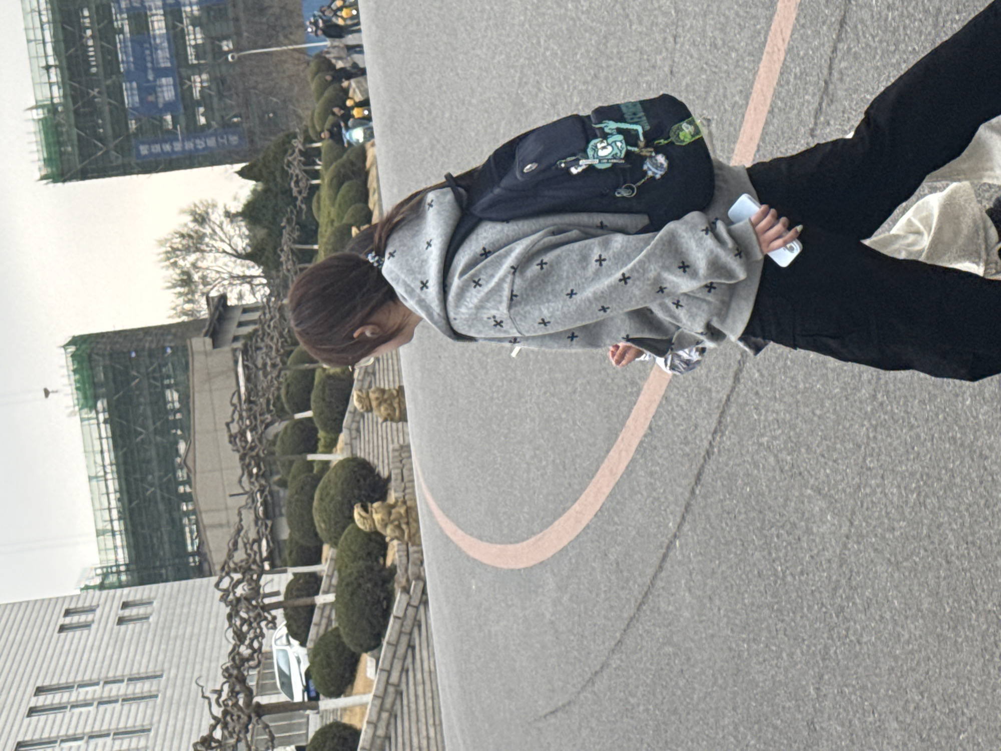A person wearing a gray hooded sweatshirt with black crosses and black pants stands on a gray asphalt surface with a peach-colored painted line. The person carries a navy backpack with green embellishments and holds a white cell phone in their right hand. A silver object is visible in their left hand. The person has long brown hair with a hair tie. The background features green round bushes, white buildings with windows, trees, construction cranes, and other people. The location is consistent with an urban area with manicured landscaping, possibly in Zhongtun, China.