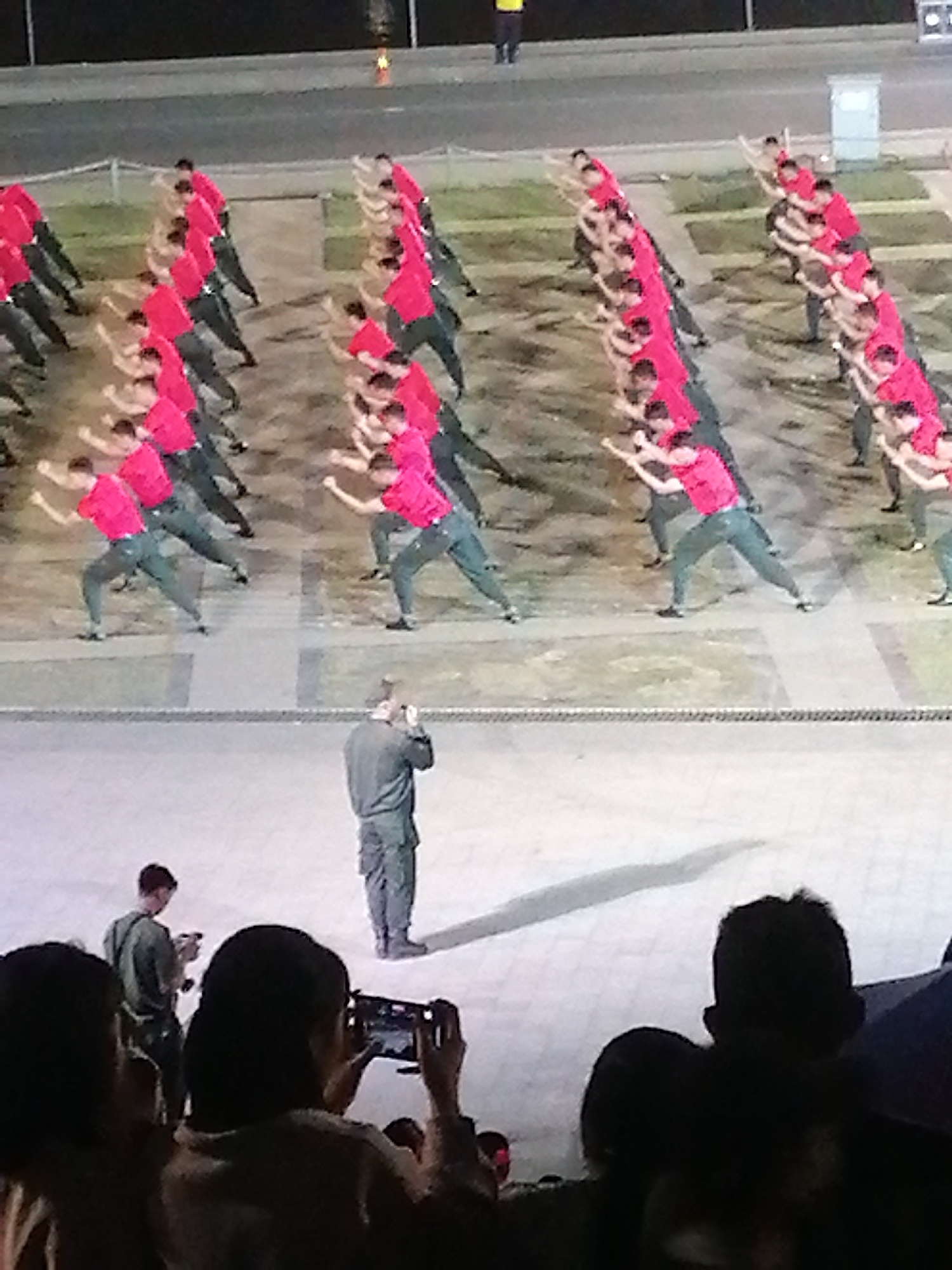 The image captures an outdoor performance in Da Lat, Vietnam, where a large group of individuals in bright red tops and dark pants are executing a synchronized routine, possibly a martial arts or drill demonstration. They are arranged in multiple rows, facing towards the camera, with their arms extended in unison. In the foreground, a person in a dark military-style uniform stands facing away from the camera, observing the performance. Further to the left, a group of spectators, partially visible and silhouetted, are present, with one individual holding up a smartphone to record the event. The background shows an elevated, dimly lit area, suggesting a nighttime or evening performance setting. The ground appears to be paved, with some grassy areas visible behind the performers, characteristic of outdoor venues or public spaces in Da Lat.