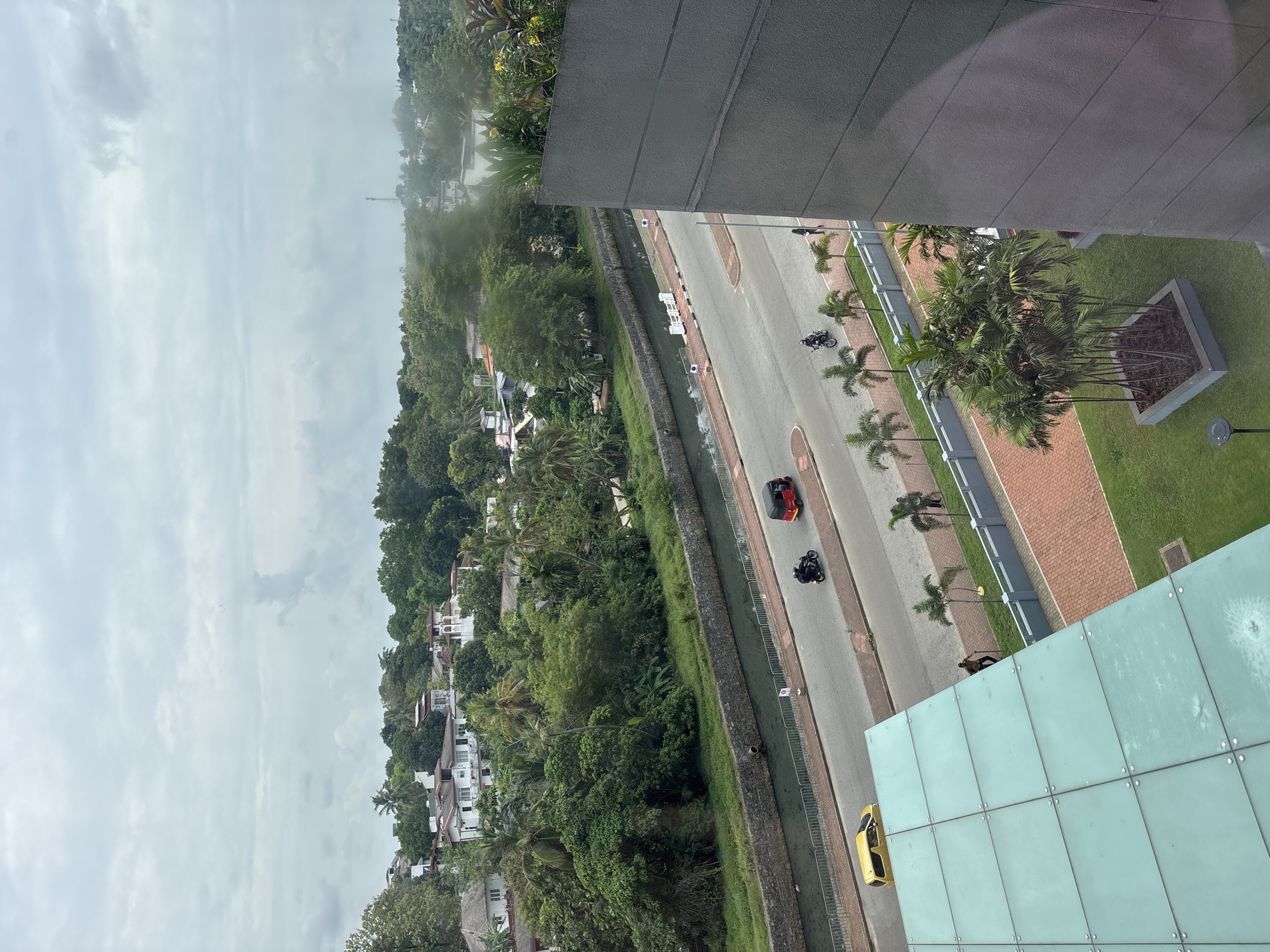 The image presents a street scene in Battaramulla, Sri Lanka, taken from an elevated perspective. A road runs through the center of the frame, bordered by a canal on one side and a green landscape with palm trees on the other. The environment includes urban elements like buildings and pavements, alongside dense vegetation, with trees and houses visible in the background. Several vehicles, including a red car, a black motorcycle, and a yellow vehicle (likely a tuk-tuk), are seen on the road. A figure is visible near the yellow vehicle, potentially interacting with it. The weather appears overcast, indicated by the cloudy sky.