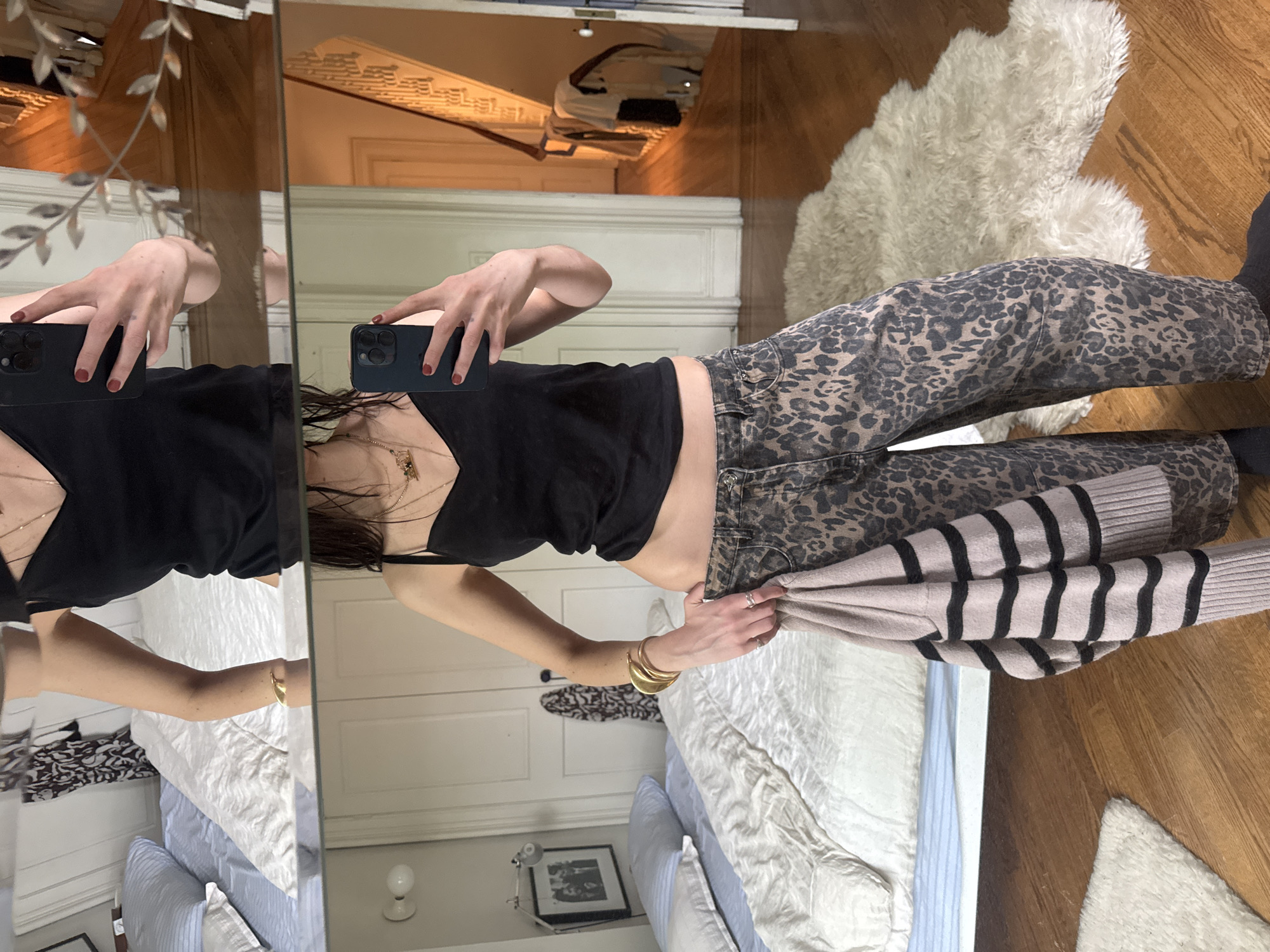 The image captures a mirror selfie taken by a person in what appears to be a bedroom. The person, whose face is not clearly visible, is holding a dark-colored smartphone with a prominent camera module in their right hand, angled to capture their reflection. They are wearing a black V-neck crop tank top, showcasing a gold layered necklace, and high-waisted pants with a brown and black leopard print. Their nails are painted red, and they wear several gold bangles on their right wrist and a ring on their left hand.
In their left hand, the person is holding a light pink or beige sweater with horizontal black stripes. The background visible in the mirror's reflection includes a wooden floor, a white fluffy rug, and a bed with white and blue striped bedding and pillows. A white paneled headboard or wall is also reflected. To the left, a white side table with a lamp and a framed picture is visible. Above, the ceiling appears to have decorative molding, and a metallic vine embellishment is attached to the mirror's frame. The overall setting suggests a private residence, potentially a stylish apartment, aligning with the given context of New York, USA. No visible text is discernible in the image.