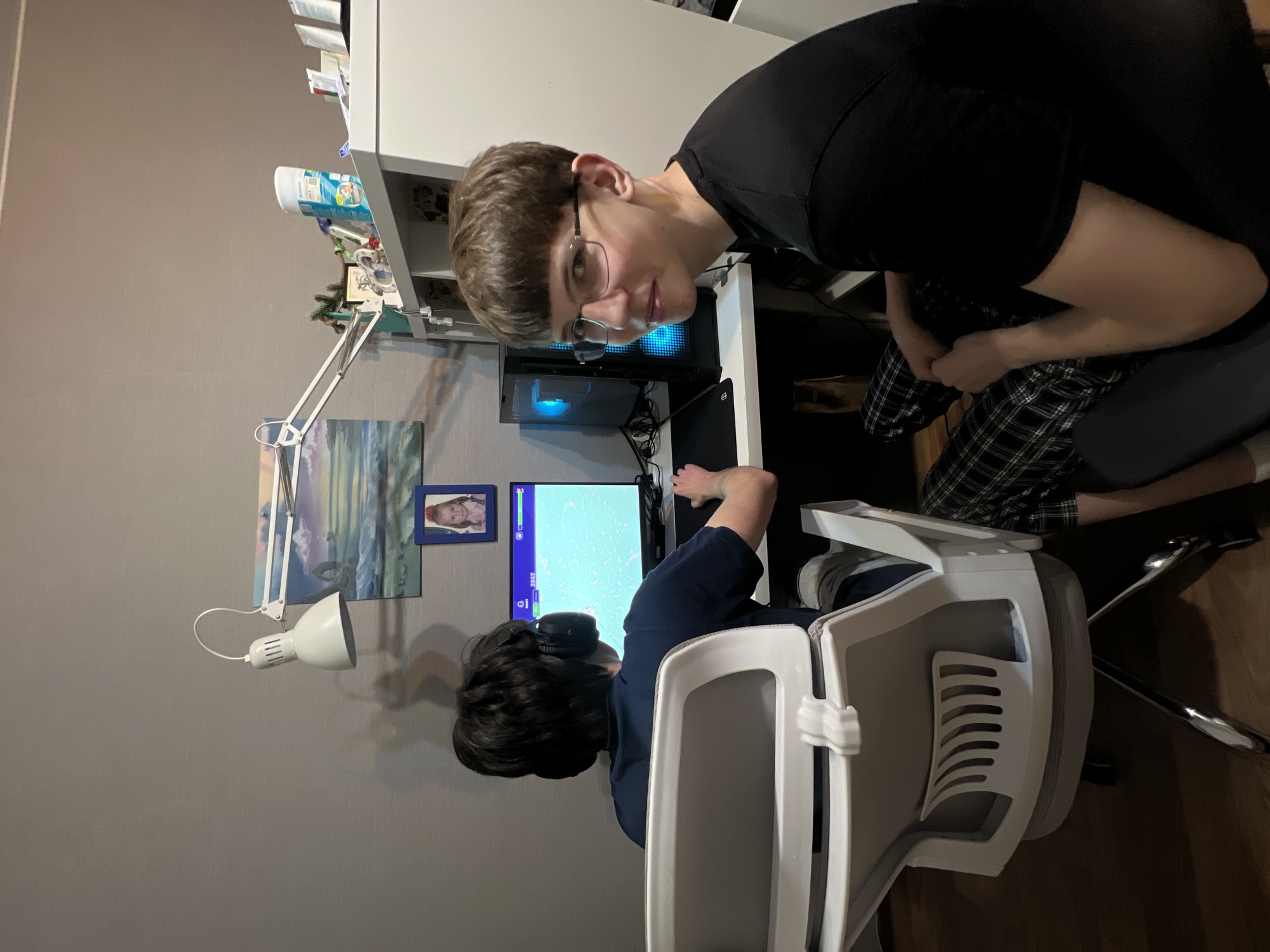 Two young men are in a room, likely a home office. One is sitting at a desk, facing a computer monitor displaying a video game interface. He is wearing a dark blue shirt and headphones. The other man is leaning over the desk, looking directly at the camera. He is wearing a black t-shirt and glasses. The desk has a white lamp and several decorative items. The room has light grey walls. There is no information to suggest the photo was taken in Dzerzhinsky, Russia.