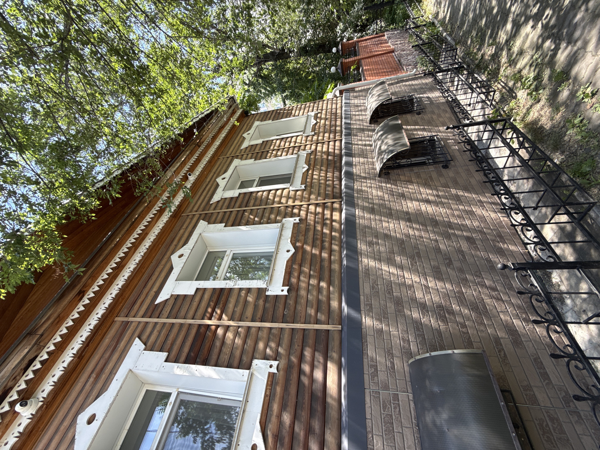 The image shows the exterior of a two-story wooden house in Khabarovsk, Russia. The house has a traditional Russian architectural style with ornate white trim around the windows and under the eaves. The lower story of the house is clad in brown brick. A black metal fence runs along the sidewalk in front of the house. Trees with green leaves provide shade over the house and the surrounding area. The sky is blue and clear. The image is taken from a low angle, looking up at the house.