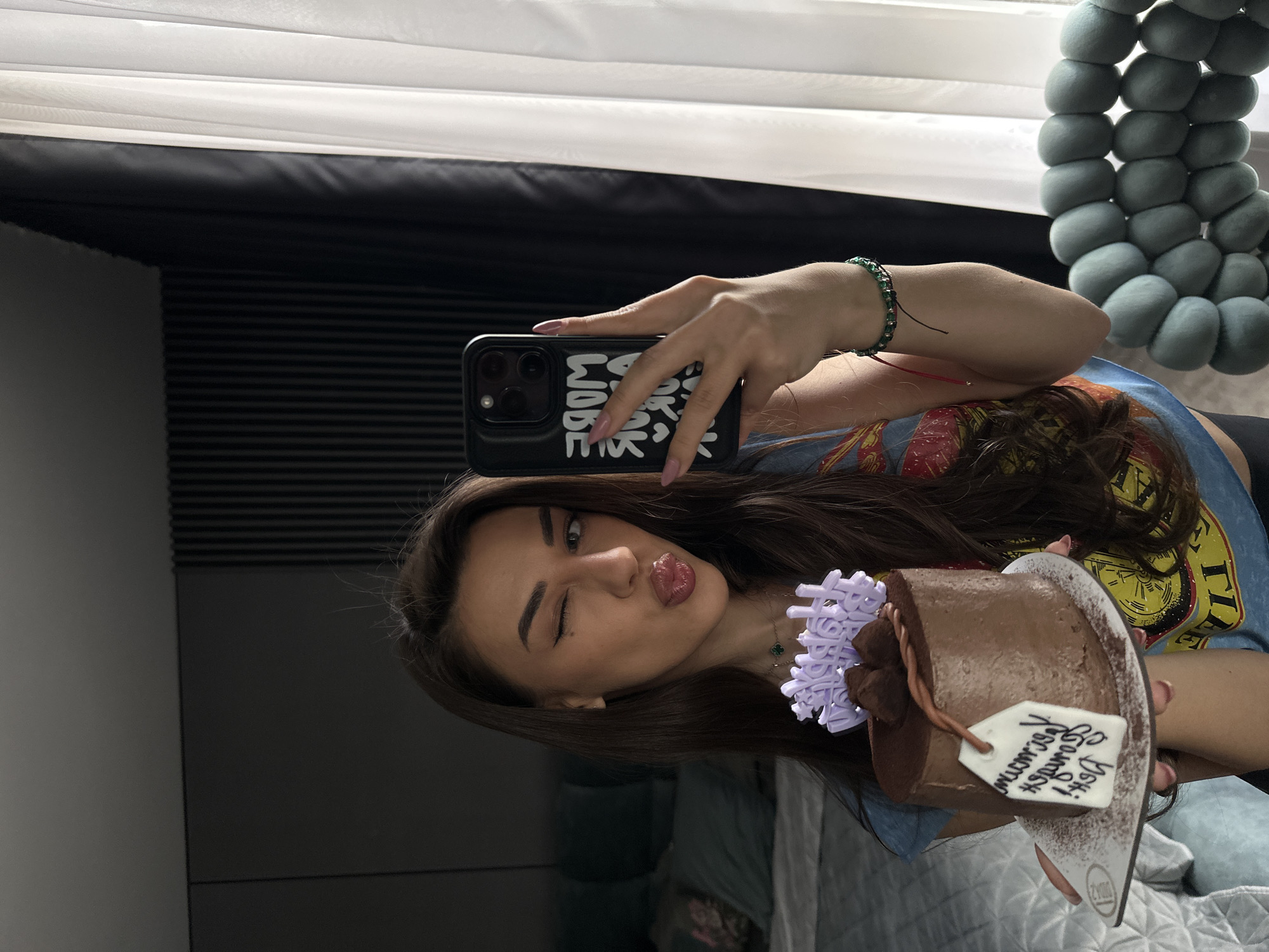 A young woman is captured in a selfie, presumably taken in a mirror, showing her from the chest up. She has long, dark hair and is winking with her left eye while puckering her lips. She is wearing a blue t-shirt with a colorful graphic print, a green beaded bracelet on her right wrist, and a small necklace featuring a four-leaf clover charm.
In her right hand, she holds a black smartphone with a case adorned with white, graffiti-style text that partially reads "WORK WIDE". With her left hand, she presents a small, round chocolate cake. The cake is decorated with a light purple, intricately cut ornament, and a white, tag-shaped piece of icing. Handwritten in black icing on this tag is the Bulgarian phrase "За най-добрата сестра," which translates to "For the best sister."
The background indicates an indoor setting, likely a home. White window blinds or curtains are visible at the top, suggesting natural light. To the left, there's a dark wall with horizontal ribbed paneling, possibly acoustic or decorative. The reflective surface of the mirror shows distorted elements of the room, including what appears to be a grey, textured decorative object made of connected felt balls on the right and hints of grey bedding or upholstery. The scene evokes a celebratory and playful mood, likely marking a birthday occasion.