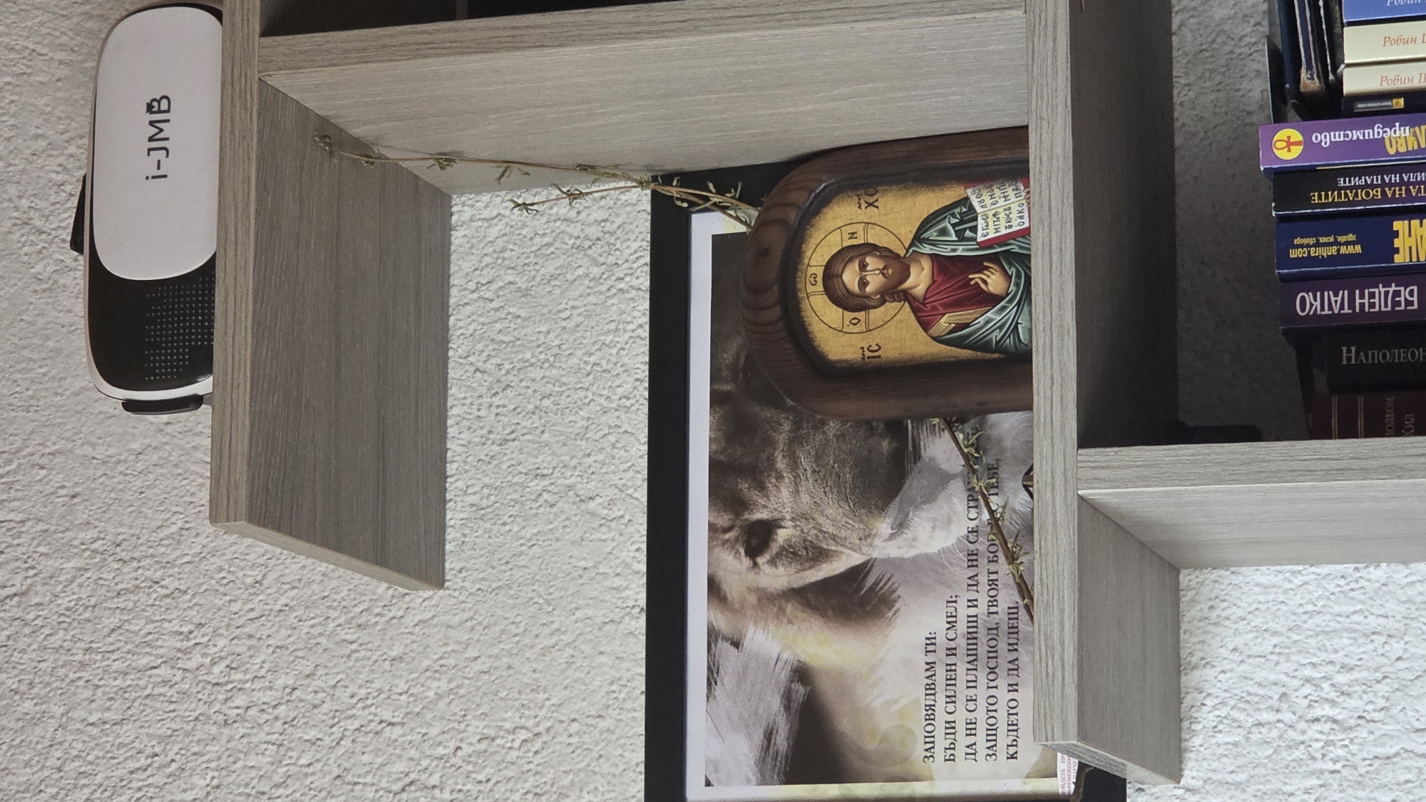 The image displays a section of a light-colored wooden bookshelf against a textured white wall, captured from a slightly tilted perspective.
On the top left, a white and black VR headset, labeled "i-JMB," is suspended from the underside of a shelf. A thin, dried plant stem or vine is draped across the top shelf, extending towards the right.
The middle shelf holds a religious icon of Jesus Christ, featuring a wooden frame and Cyrillic/Greek lettering including "IC XC" (Jesus Christ monogram) and "O N". Leaning next to the icon is a book or poster showcasing a close-up image of a cat or lion. Overlaying this image is Bulgarian text that translates to a motivational or biblical passage: "I command you: Be strong and courageous; Do not be afraid or fear, because the Lord, your God, wherever you go."
To the right, a stack of books is visible on the shelves. Titles and authors in Bulgarian include "Робин Шарма" (Robin Sharma), "ПРЕДИМСТВО" (Advantage), "ПЪТЯ НА БОГАТИТЕ" (The Way of the Rich), "ИГРА НА ПАРИТЕ" (The Game of Money), "АНХИРА" (Anhira) with "www.anhira.com" and "здраве, успех, свобода" (health, success, freedom), and "БЕДЕН ТАТКО" (Poor Dad), likely by "НАПОЛЕОН ХИЛ" (Napoleon Hill).
The scene suggests an organized display of personal items, combining modern technology, religious artifacts, and various literature. No specific activity or event is taking place; the image is a static shot of the arrangement.